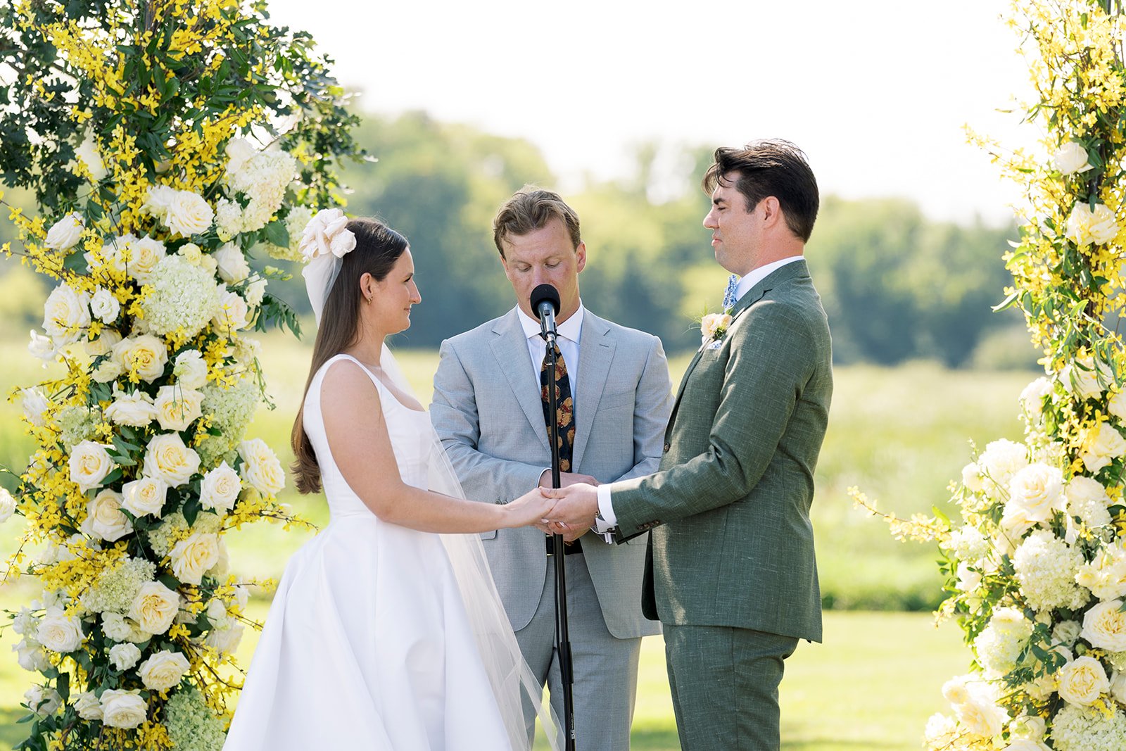 A couple getting married outdoors under a floral arch, with an officiant in a gray suit conducting the ceremony.