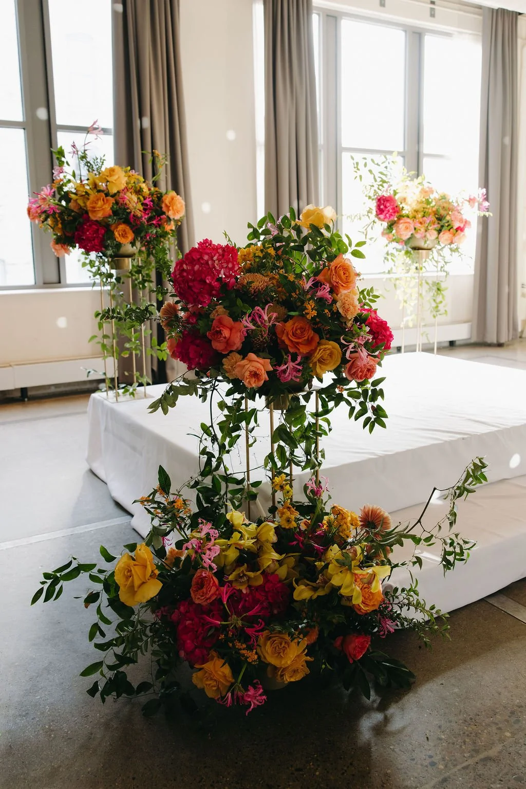 Colorful flower arrangements on stands in a bright room with large windows and curtains.