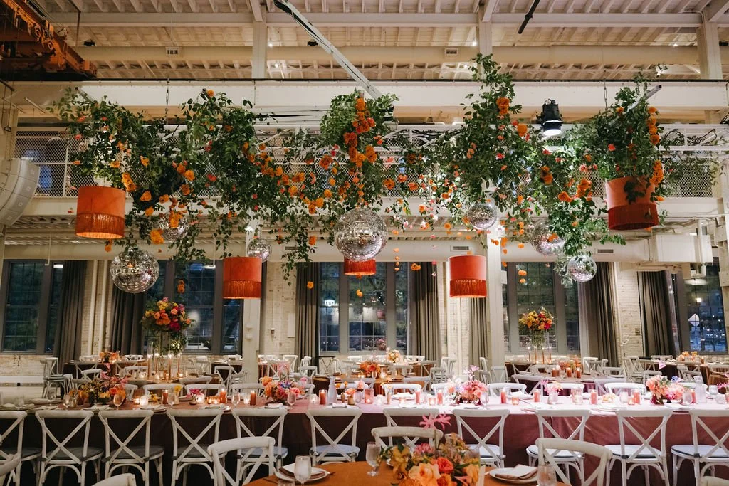 Elegant event space with floral decorations, hanging greenery, orange and gold lamps, and disco balls, featuring a long banquet table with floral centerpieces and candles, set for a celebration or wedding.