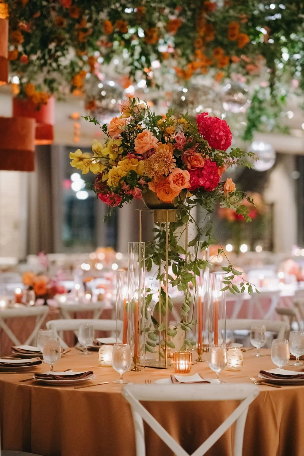 Elegant banquet table with a tall, colorful floral centerpiece, candles, orange tablecloth, and arranged dinnerware, set in a decorated venue.