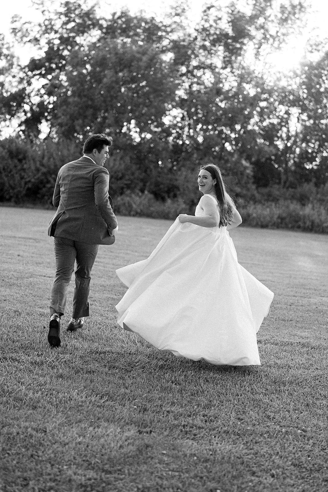 A black-and-white photo of a bride and groom outdoors on grass, smiling and dancing, with trees in the background.