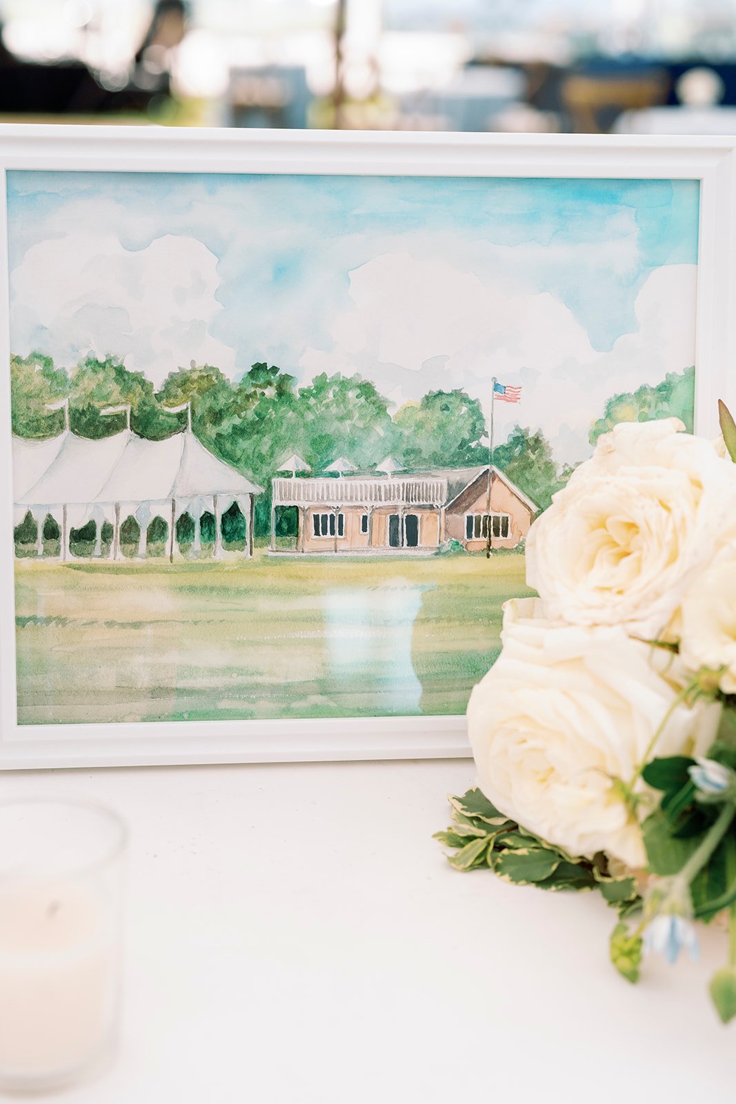 A framed watercolor painting of a large outdoor tent and building with a flag, surrounded by trees, with a grassy area in the foreground and a bouquet of white roses on the right side of the image.