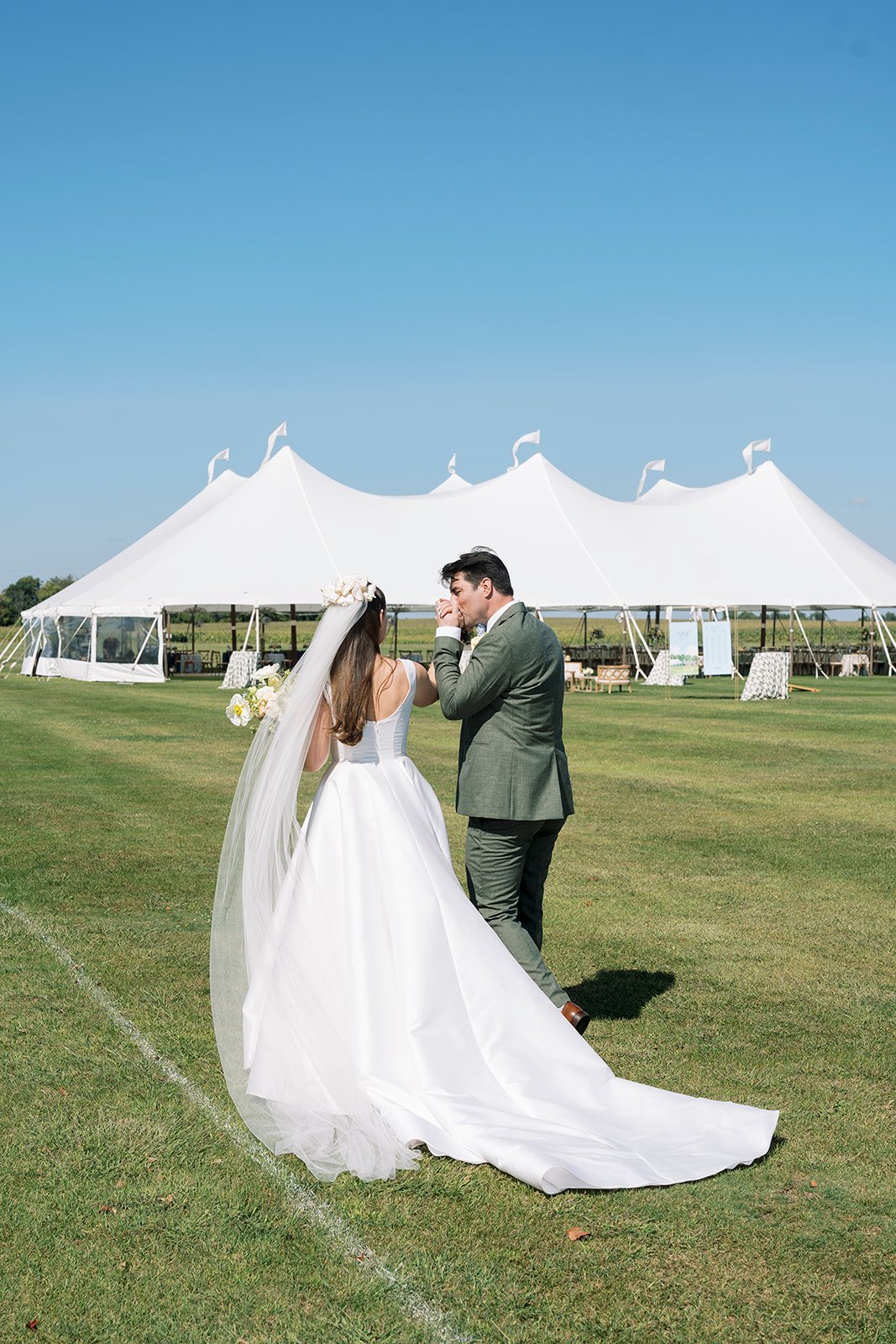 A bride and groom share a moment on her wedding day outdoors on a grassy field, with a large white tent in the background under a clear blue sky.