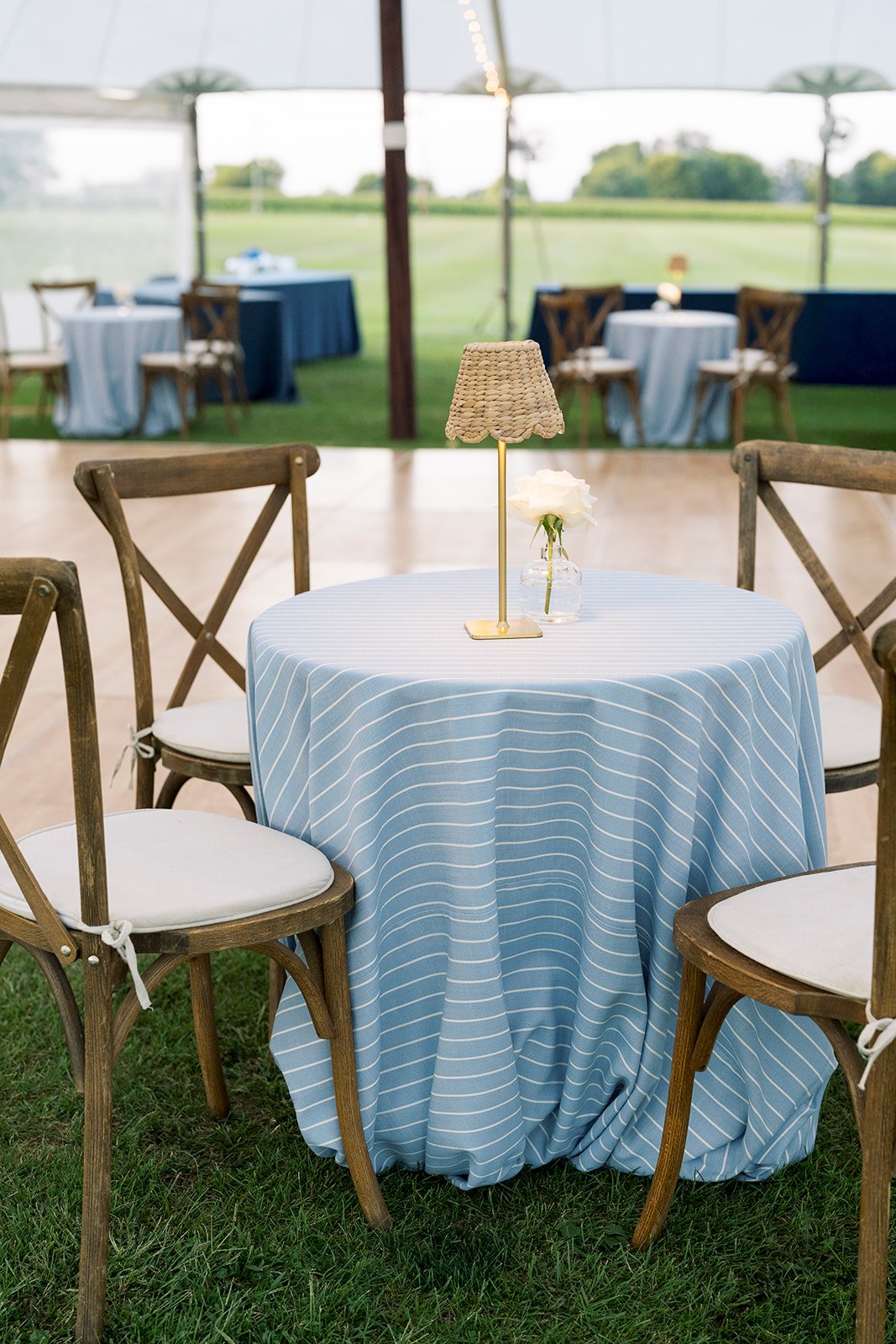 Round table with blue and white striped tablecloth, surrounded by wooden chairs with white cushions, indoors with a view of an outdoor event space with additional tables and chairs under a tent.