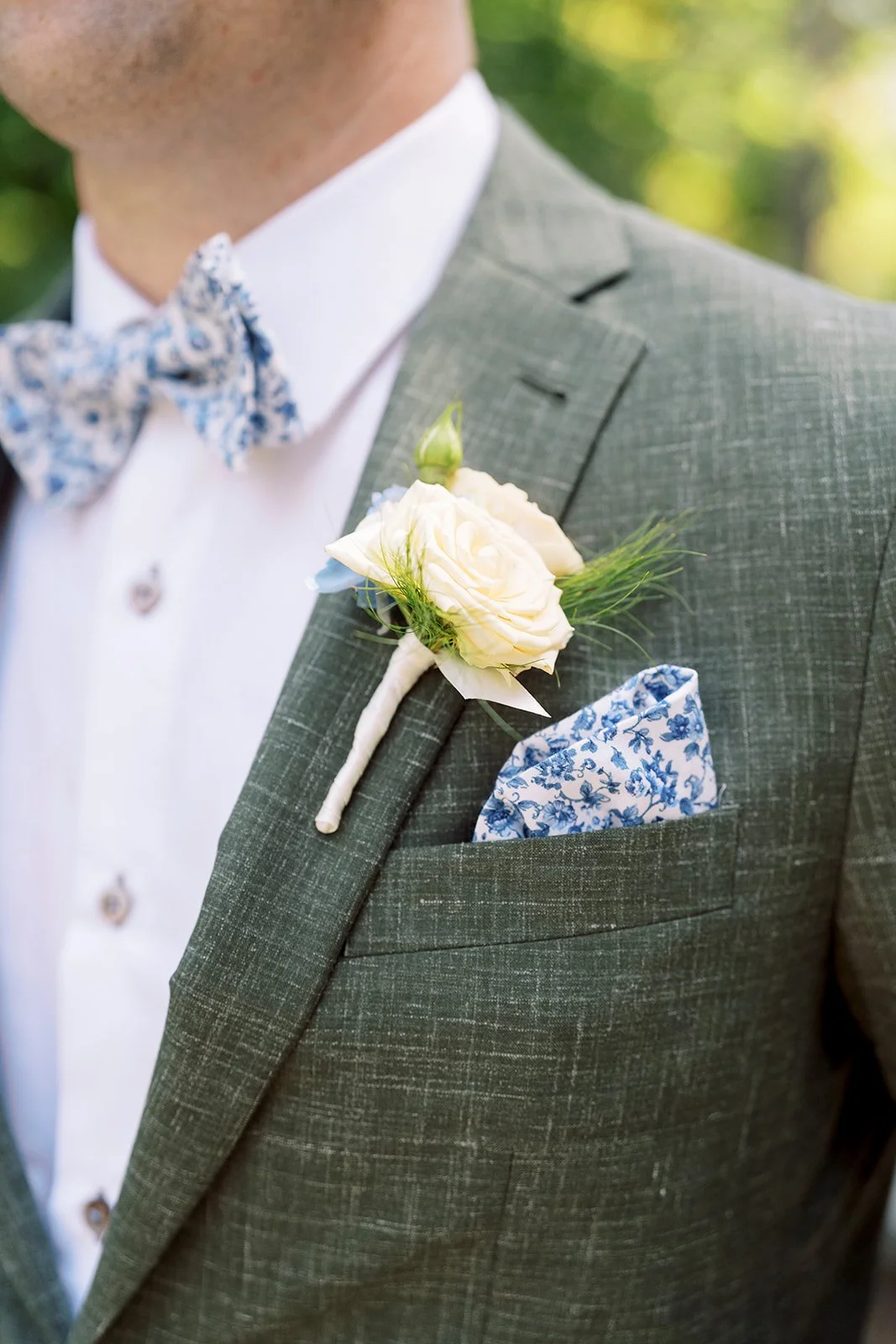 Close-up of a groom's gray suit with a white boutonniere and a blue floral pocket square, outdoors with blurred greenery in the background.