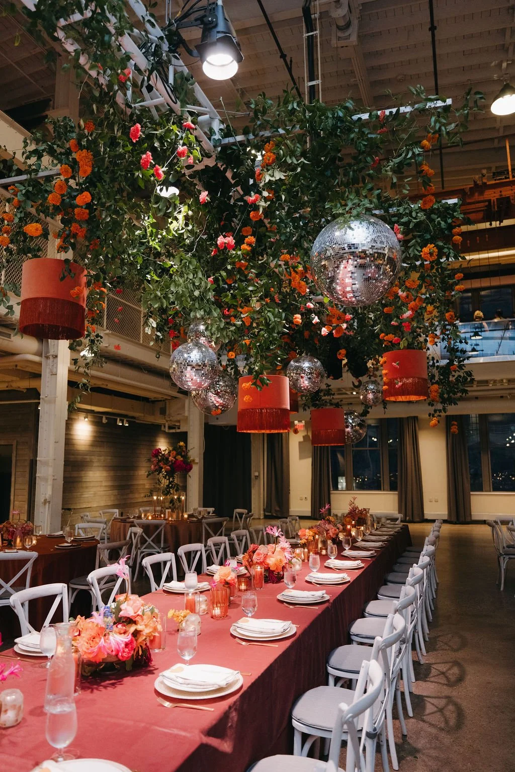 A decorated event space with a long dining table, floral centerpieces, and hanging disco balls, with a ceiling covered in greenery and orange flowers, warm lighting, and large windows at the back.