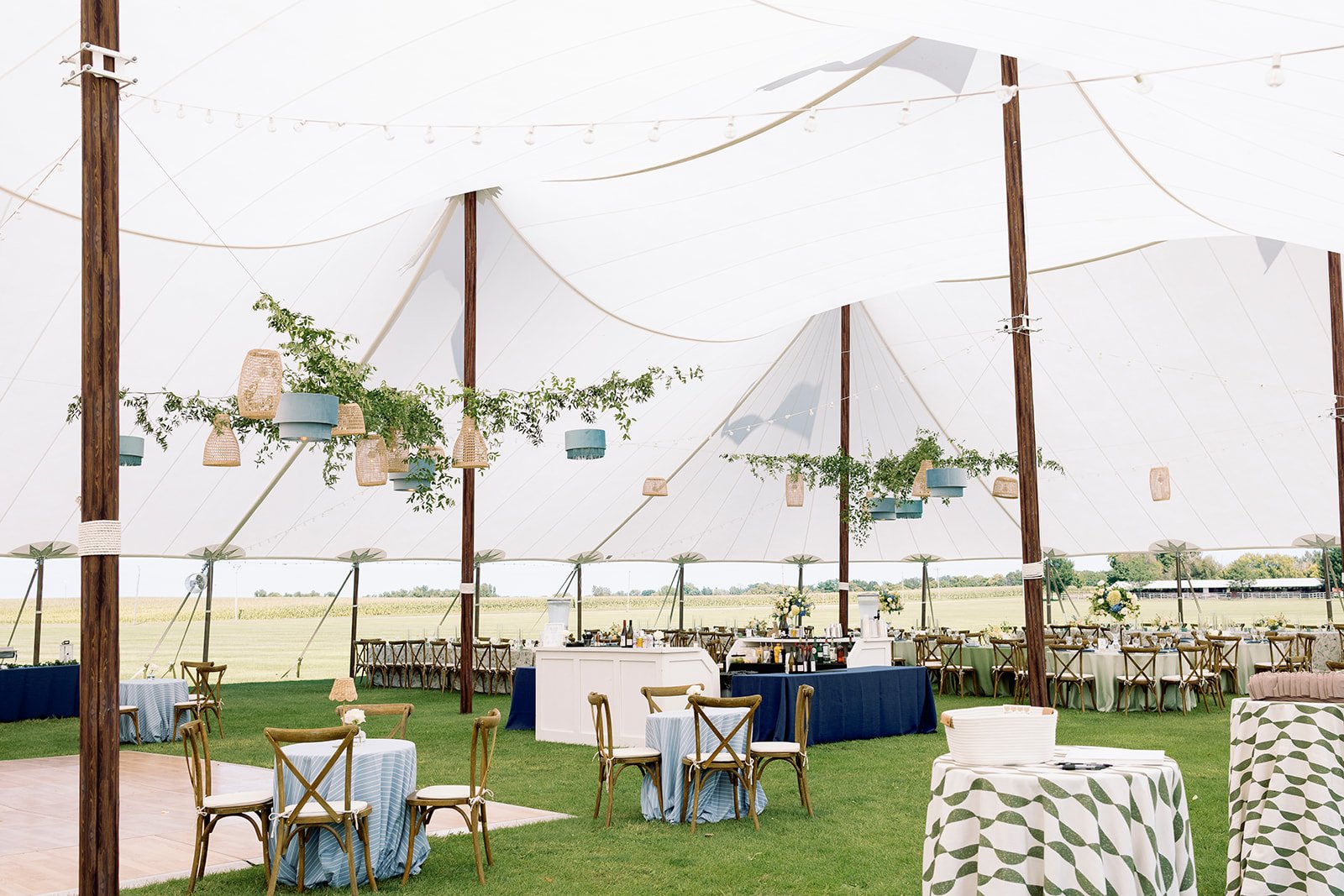 Decorated outdoor event tent with tables, chairs, and hanging lanterns, overlooking a field.