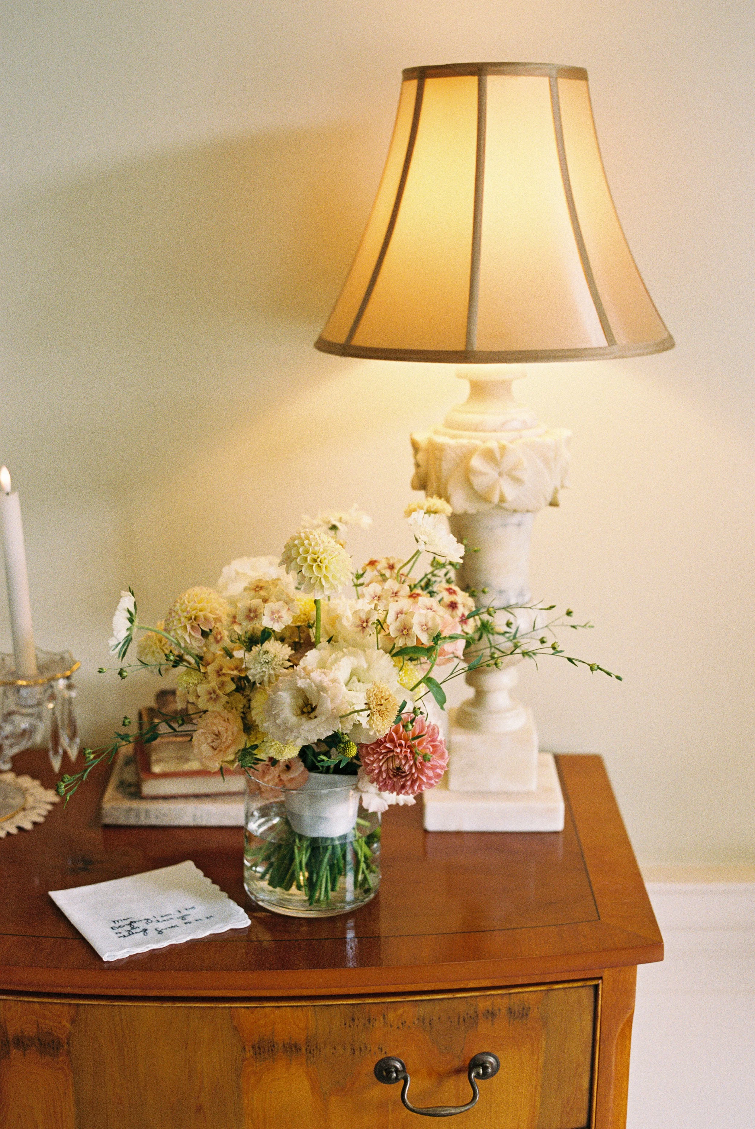 A wooden table with a floral bouquet in a glass vase, a white ceramic lamp with a beige shade, a candle, and a small cloth with handwritten notes.