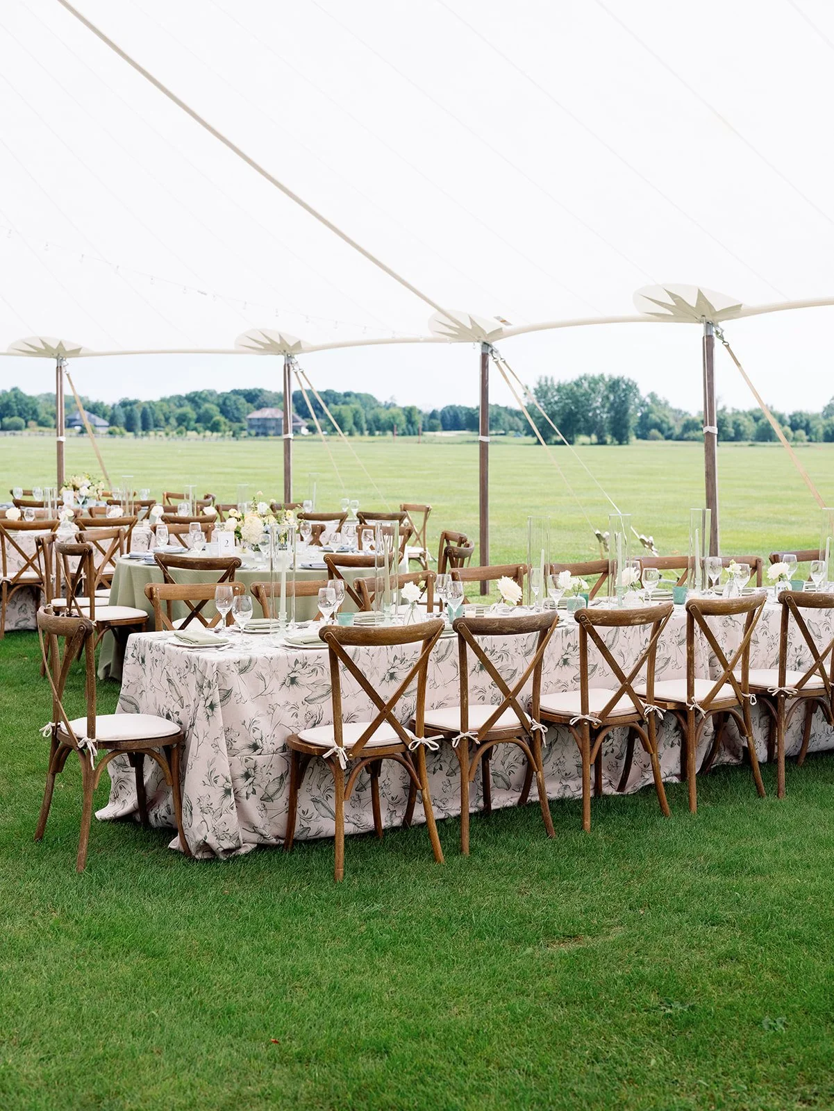 Outdoor event setup with rectangular tables covered in floral tablecloths and wooden cross-back chairs, set under a large white canopy tent in a green field with trees and houses in the distance.