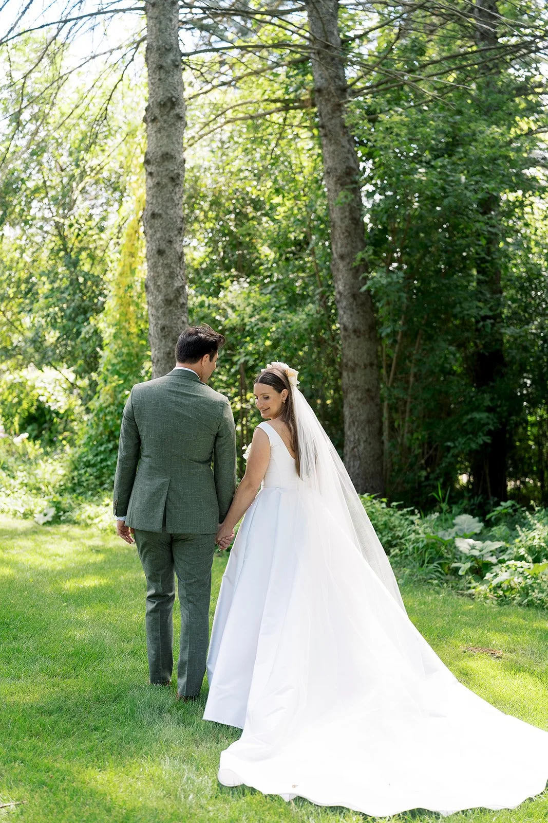 Bride and groom holding hands walking in a green wooded outdoor setting.