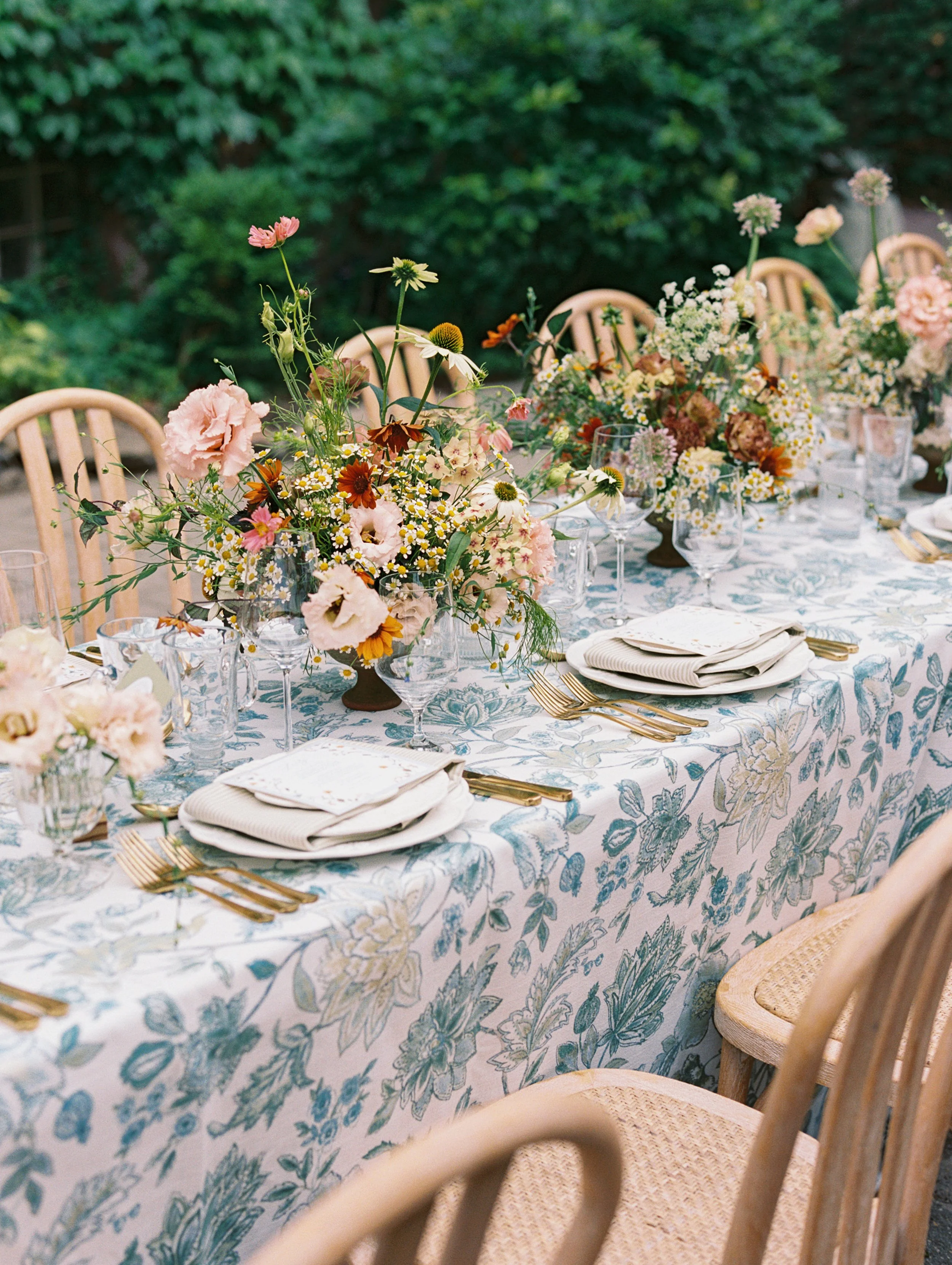 A long outdoor dining table decorated with floral centerpieces, gold cutlery, and white napkins, set in a garden with green bushes in the background.