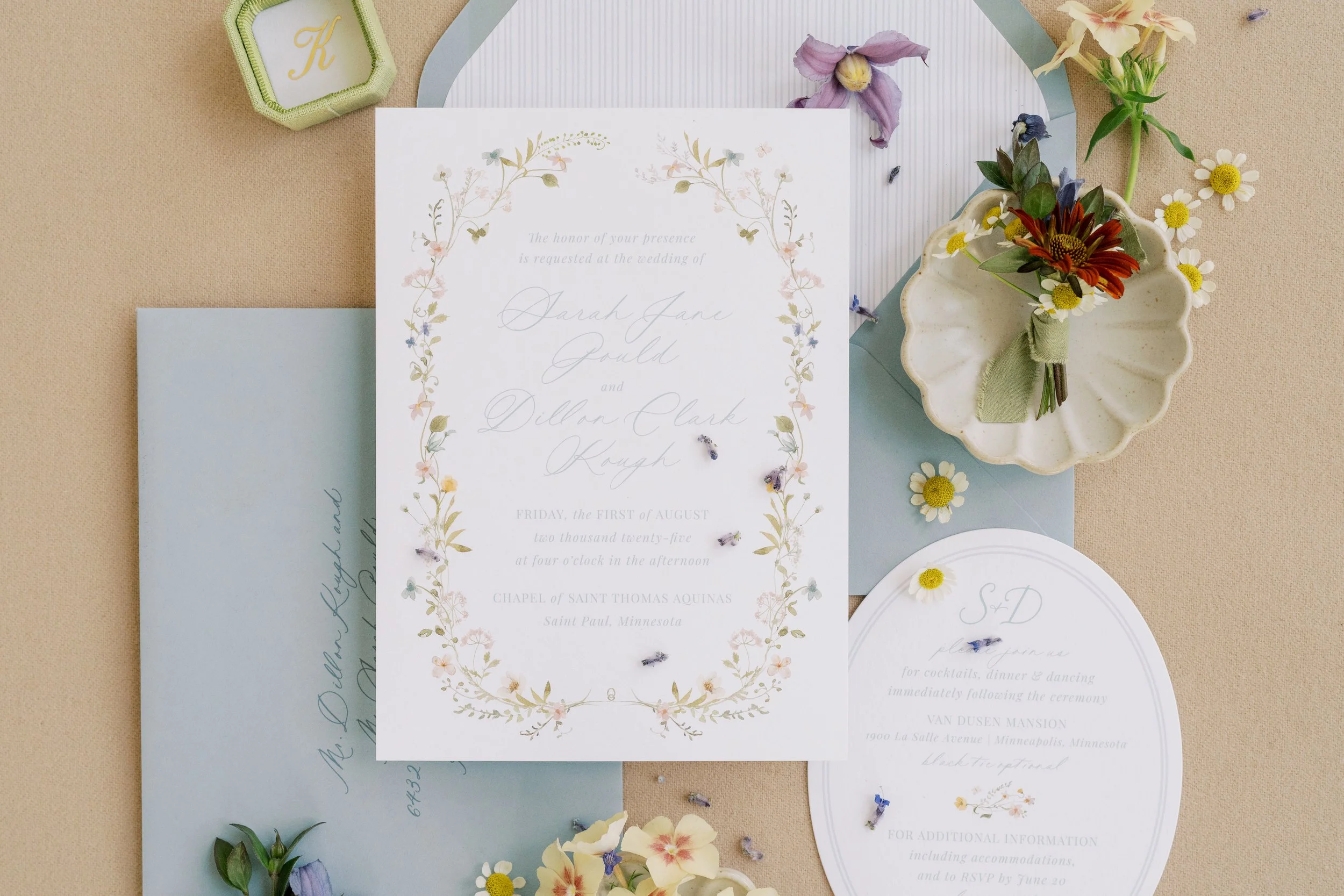 Wedding invitation suite with floral accents, including an RSVP card, a mailing envelope, and a boutonniere with daisies, placed on a beige background.