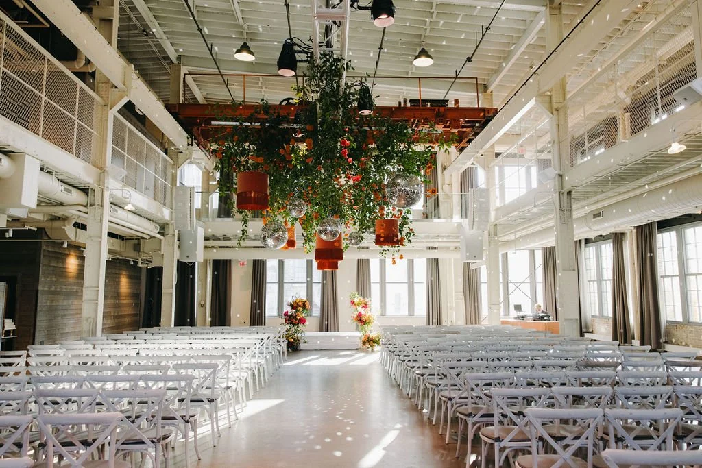 An empty indoor event space decorated for a wedding with rows of white chairs facing a floral altar and hanging floral arrangements from the ceiling.