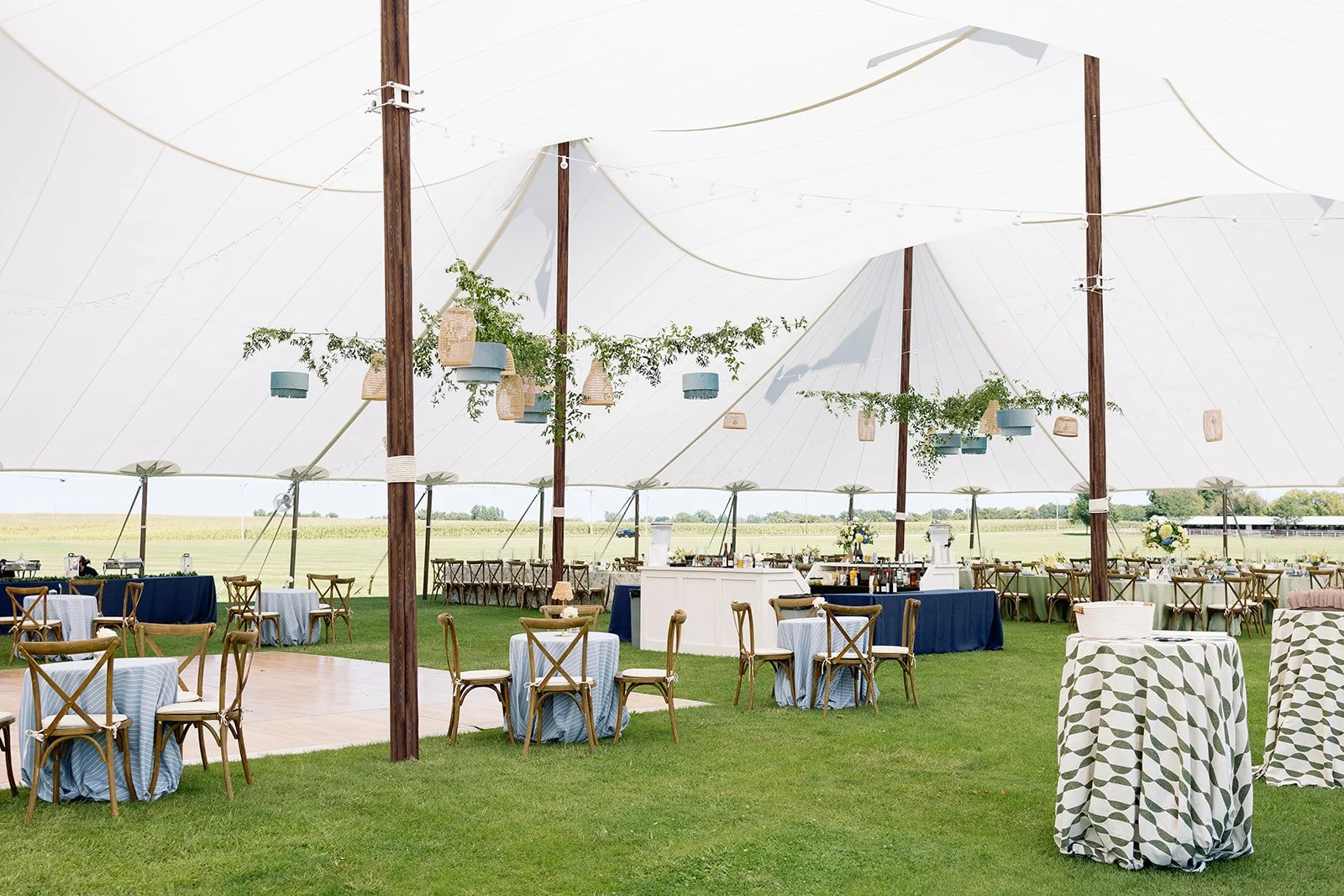 Under a large white tent with wooden supports, there are round tables with blue and white tablecloths, some with chairs and some with high-top tables covered with green and white patterned tablecloths. Hanging from the ceiling are decorated baskets and greenery, and in the background, there is a grassy open field with additional seating and a bar area.