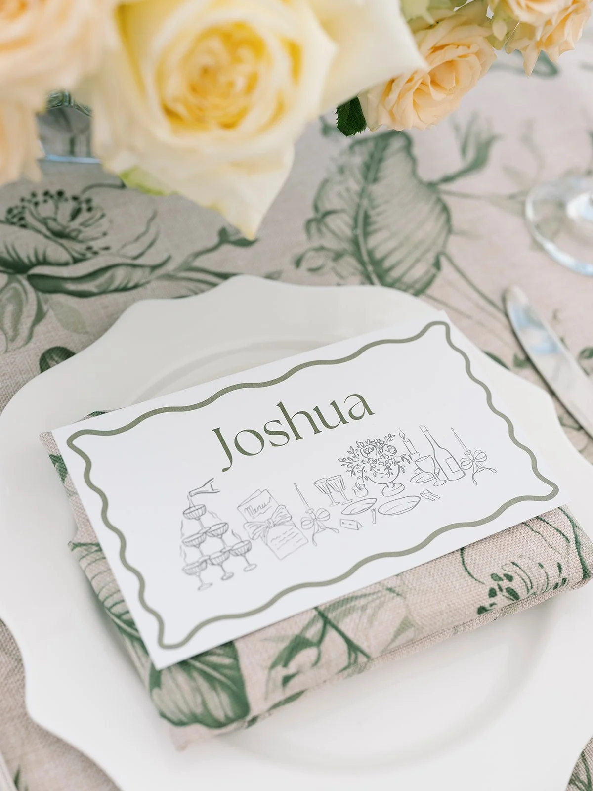 Place setting with a folded napkin, a place card with the name Joshua, and a floral centerpiece on a decorated table.