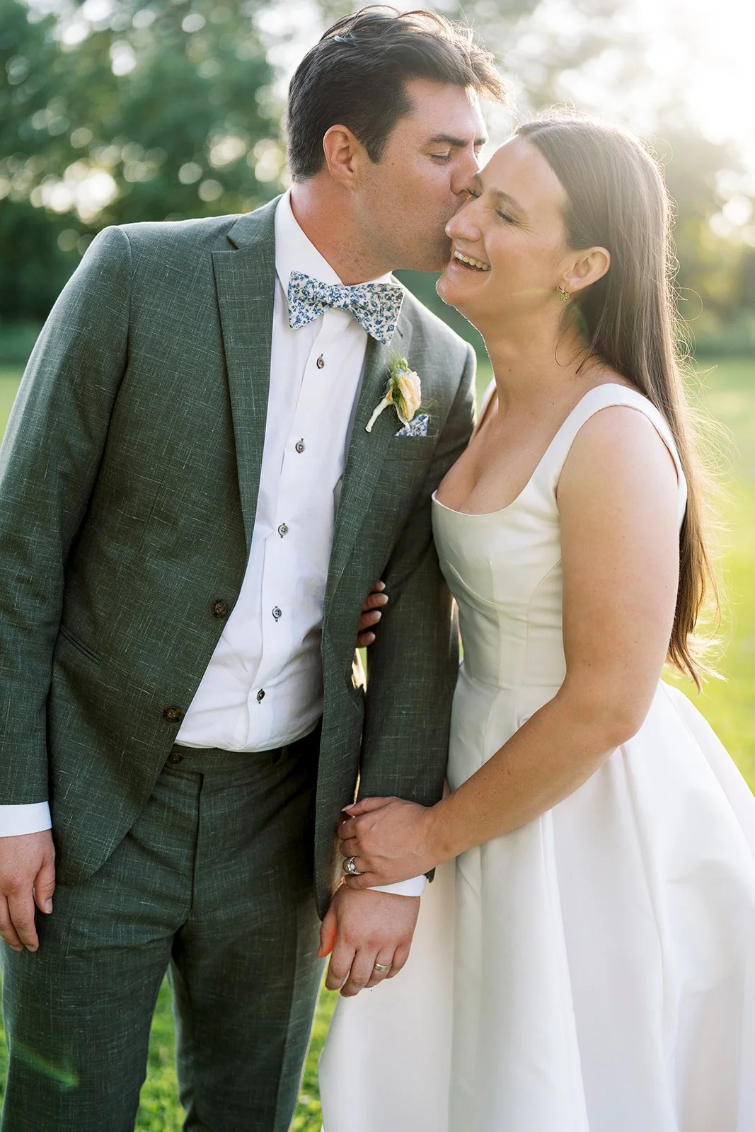 A groom in a gray suit and a bride in a white wedding dress sharing a kiss outdoors, with the groom kissing the bride on the cheek, both holding hands, in a sunny park setting.
