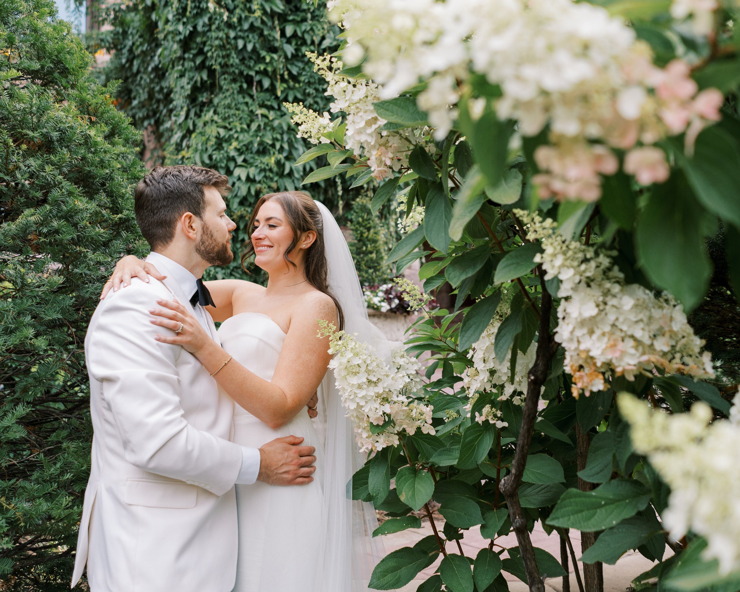 A bride and groom in wedding attire smiling and holding each other outdoors among lush green foliage and blooming white flowers.
