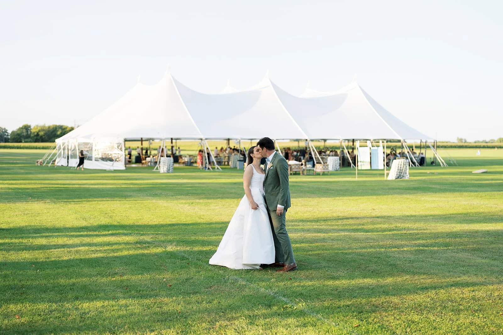 A bride and groom kissing in front of a large white wedding tent on a grassy field during daytime.