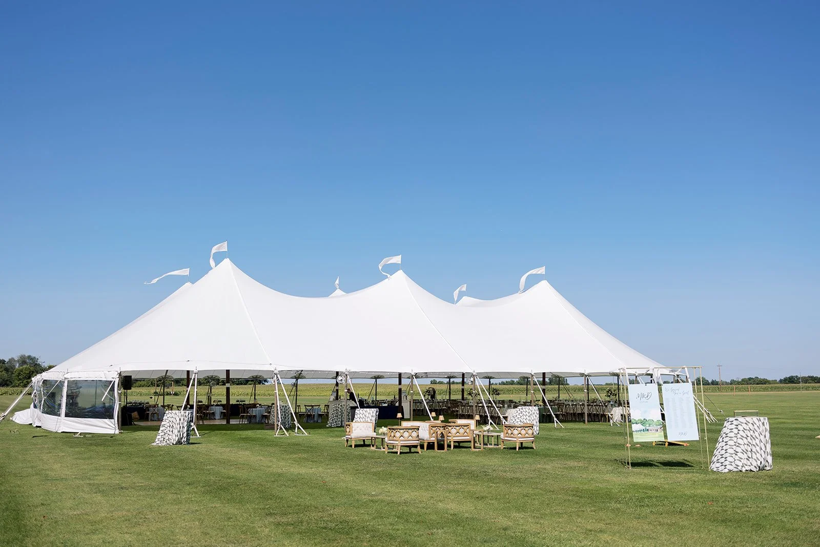 A large white event tent set up on a grassy field with clear blue sky, surrounded by outdoor furniture including chairs, tables, and artistic decor elements.