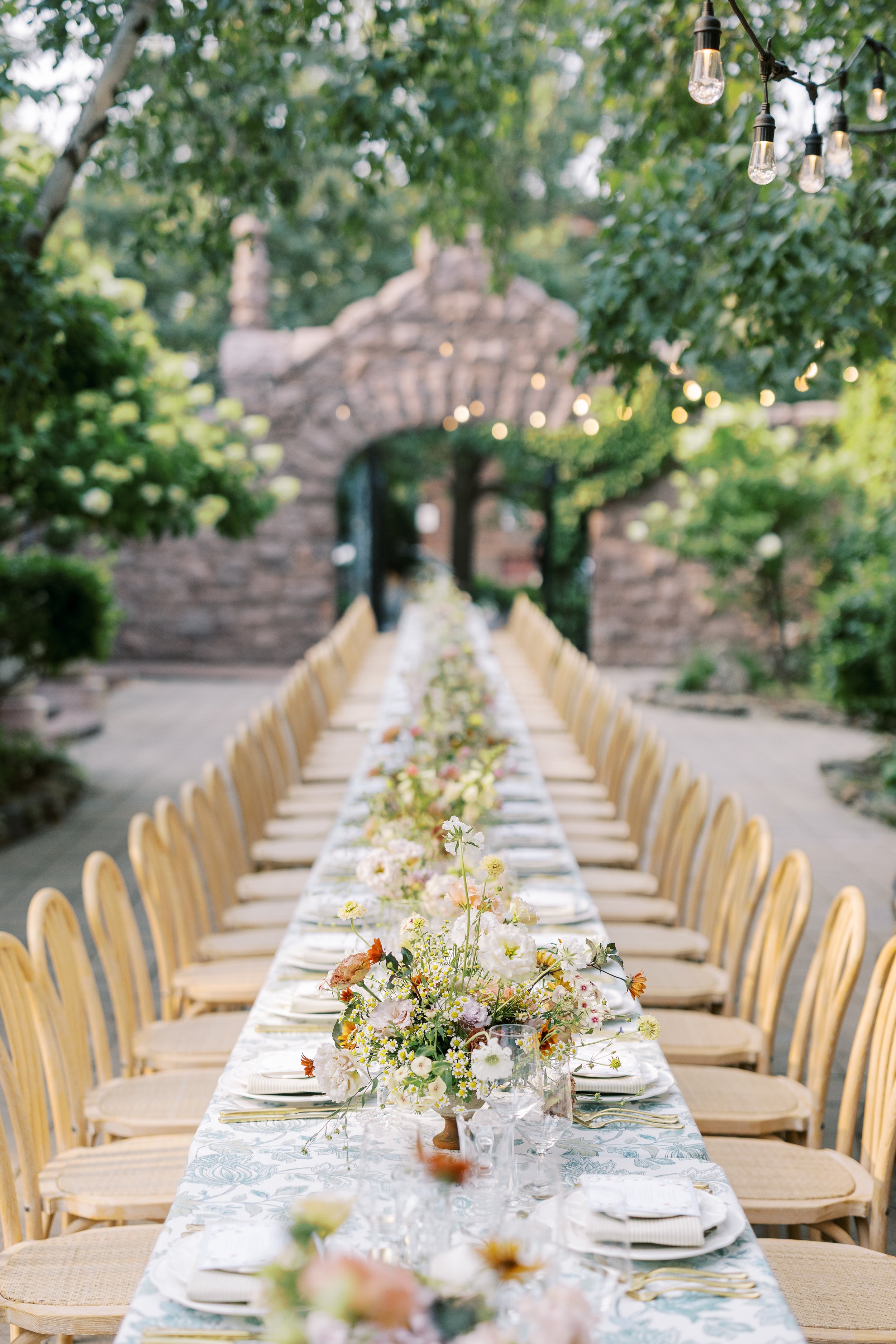 Long outdoor dining table decorated with floral centerpieces, set with white plates, gold utensils, and glasses, in a lush garden with string lights hanging overhead.