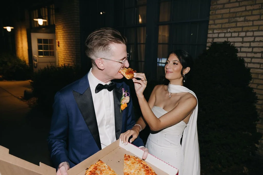 A bride feeding pizza to a groom in a tuxedo outside at night.
