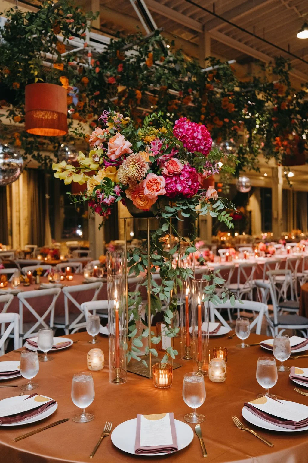 Elegant banquet table setup featuring a large floral centerpiece with pink and peach flowers, surrounded by candles and glassware in a decorated event space.