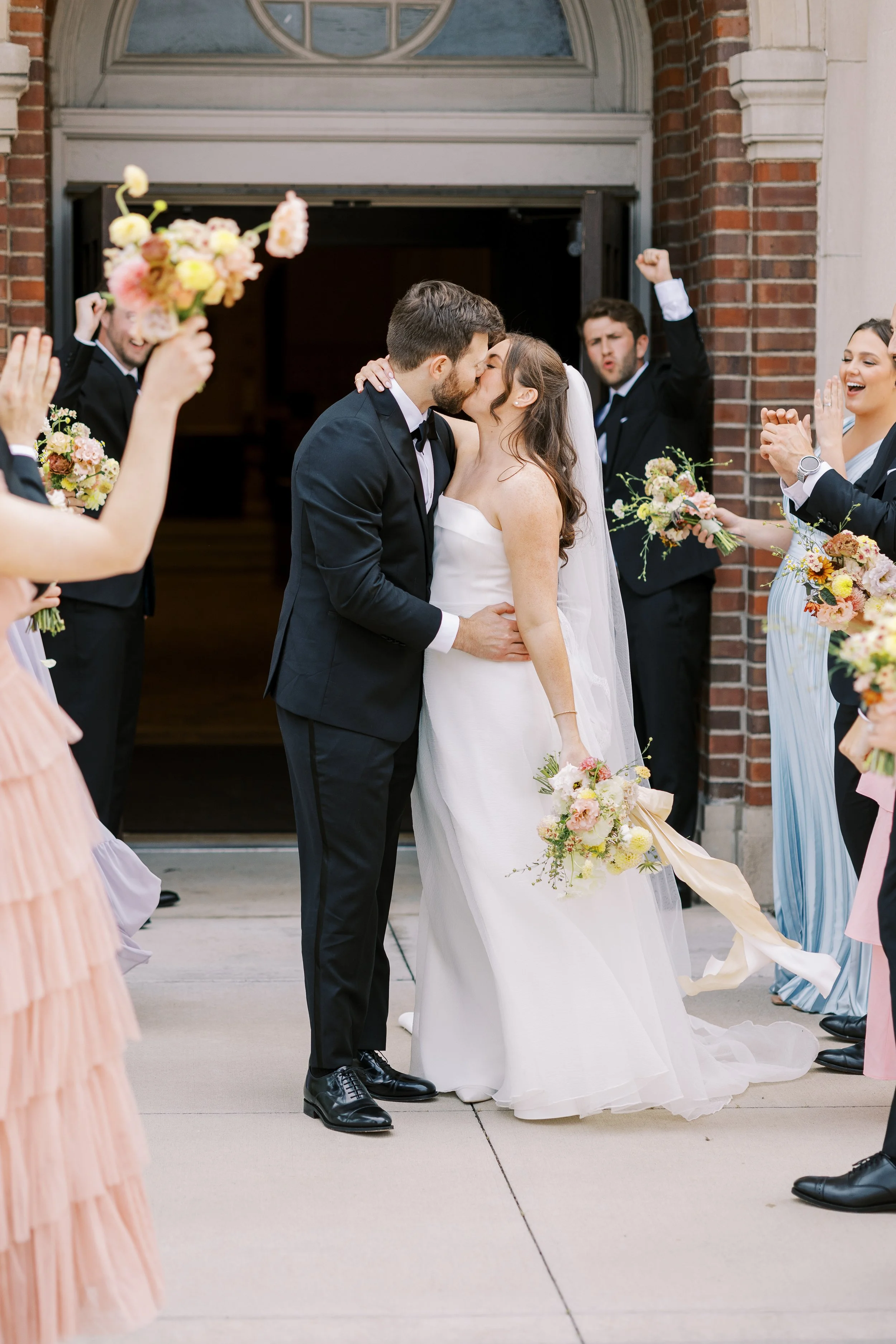 A newlywed couple sharing a kiss outside a church, surrounded by friends dressed in colorful attire, celebrating their wedding.