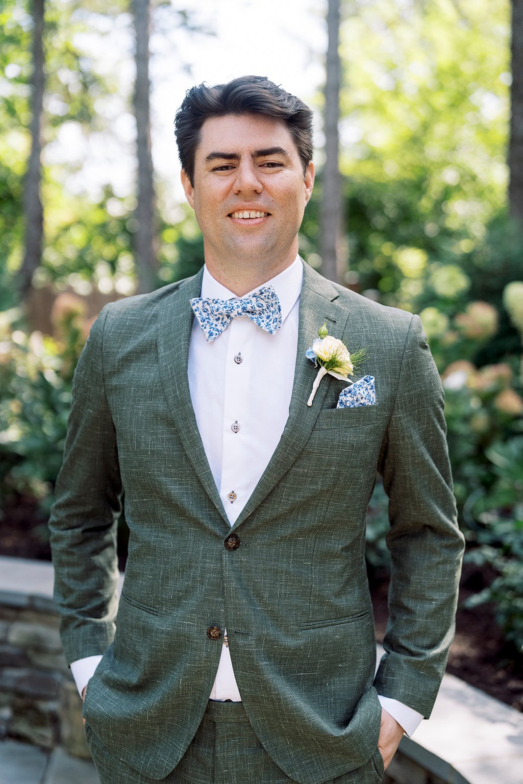 A man in a green suit with a white shirt, patterned bow tie, and matching pocket square, standing outdoors with greenery in the background.