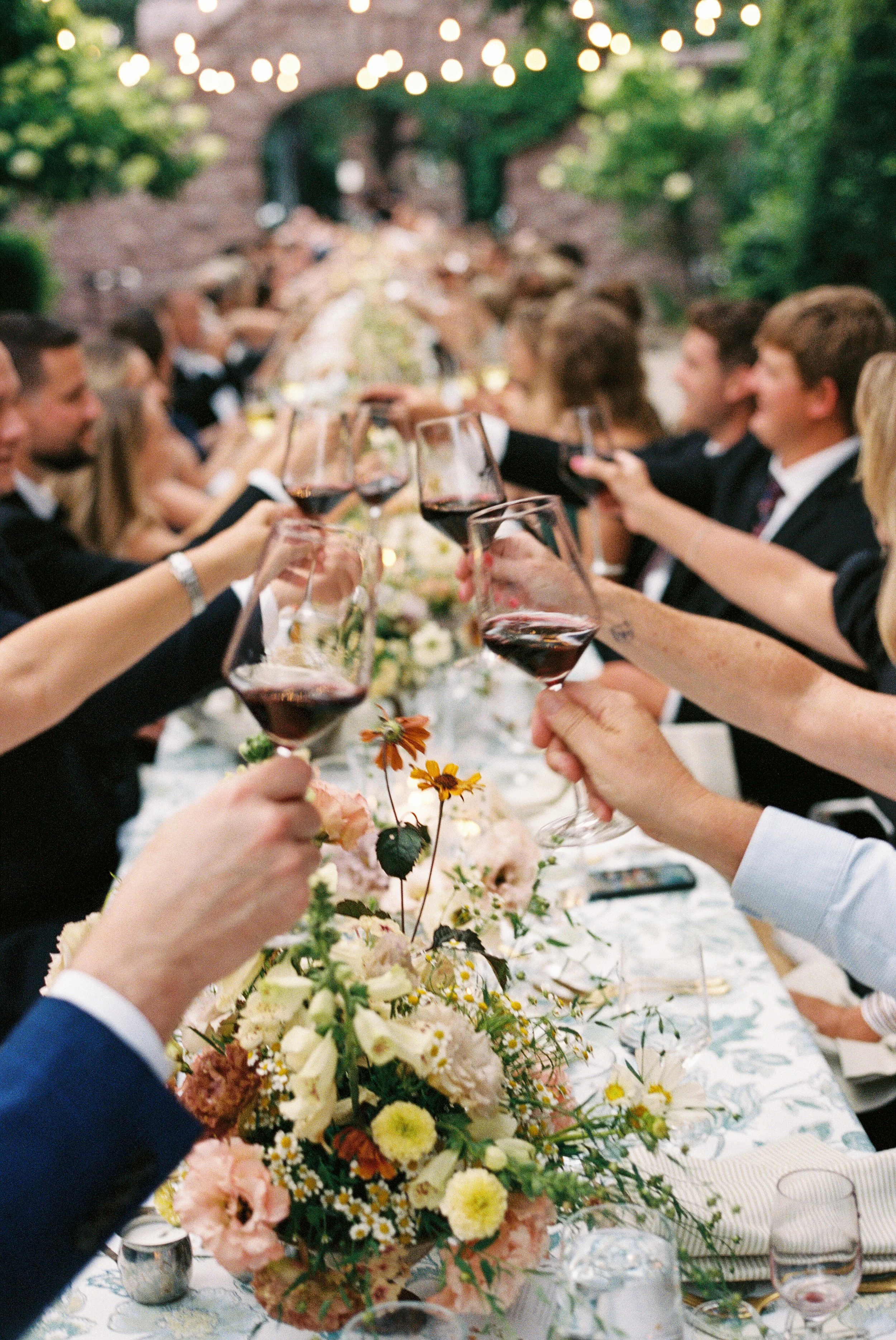 People celebrating with glasses of red wine at a long outdoor dinner table decorated with flowers, under string lights.