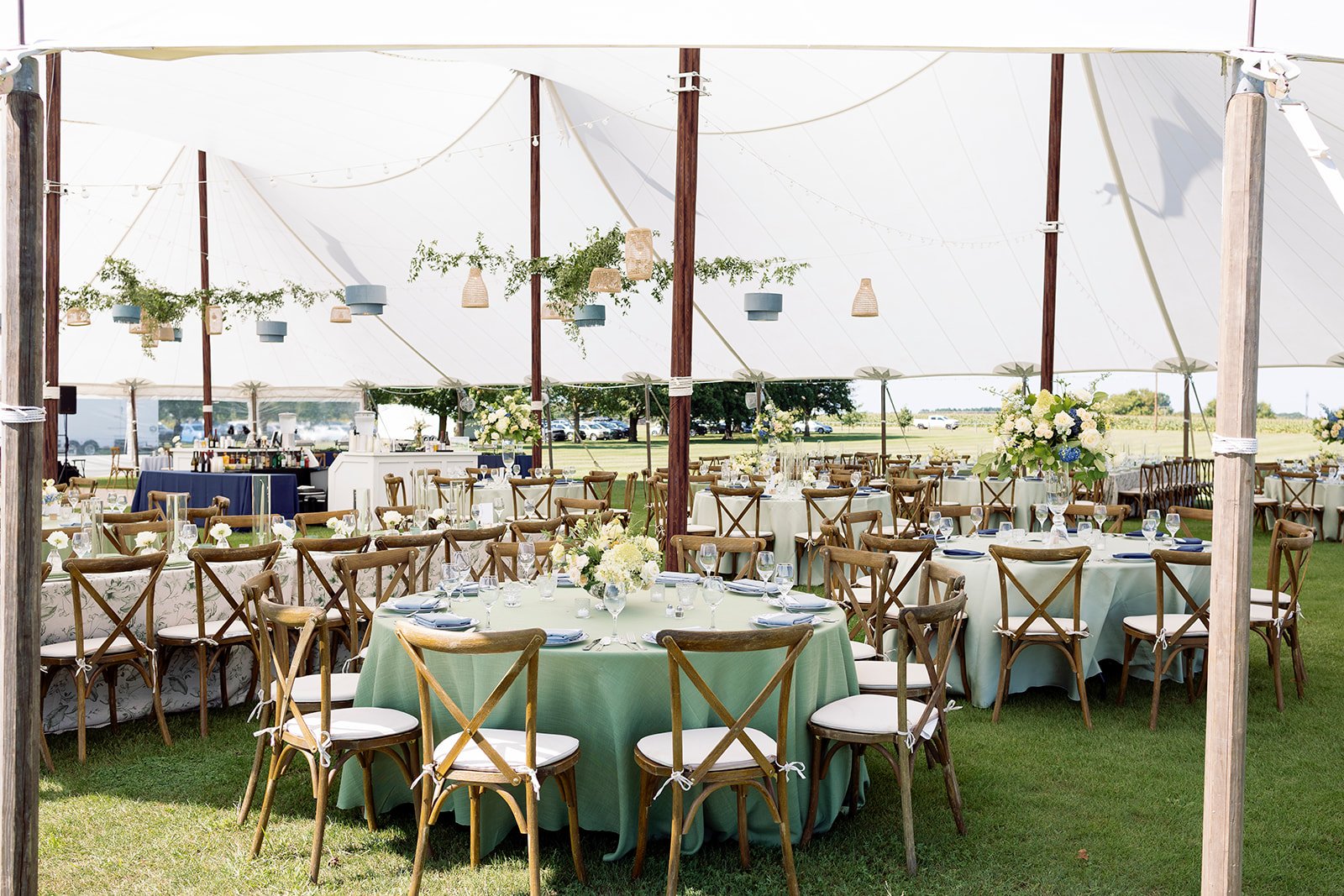 Wedding reception setup inside a large white tent with round tables decorated with white and blue floral centerpieces and wooden chairs with white cushions, on a grassy field with trees in the background.