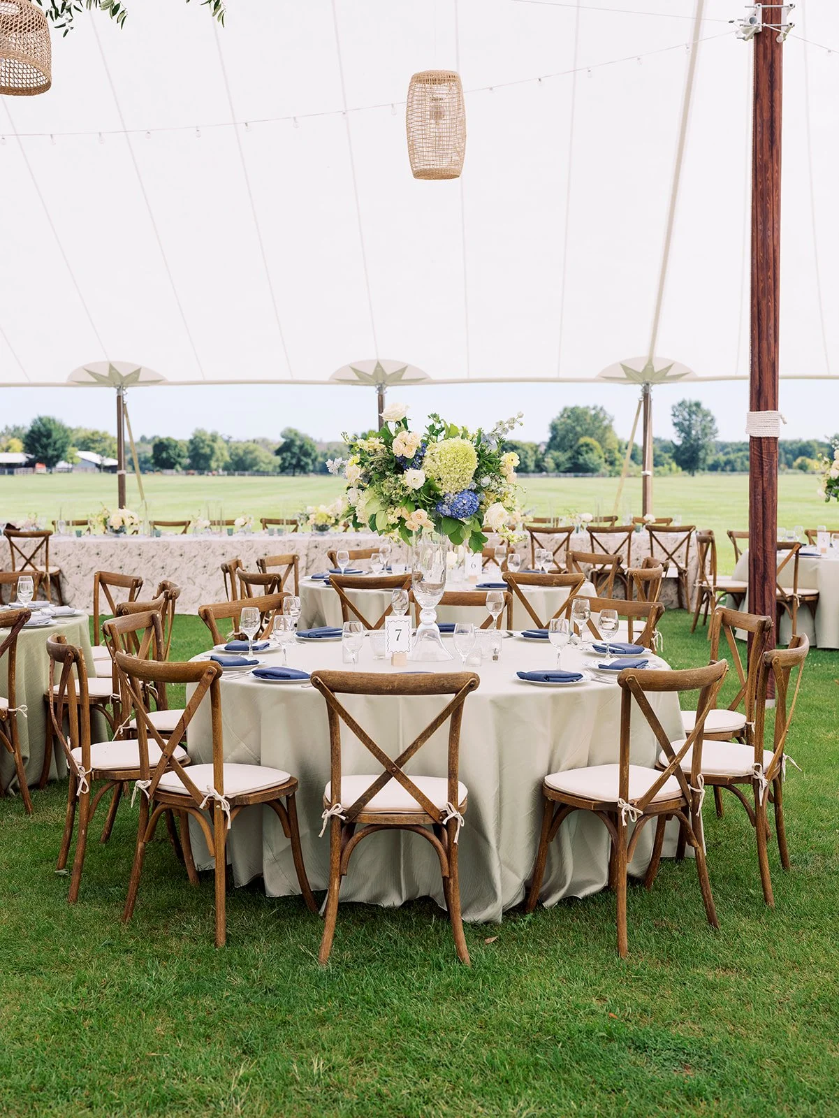 Round outdoor banquet setup with white tablecloths, wooden cross-back chairs, and a tall floral centerpiece under a white tent with string lights.