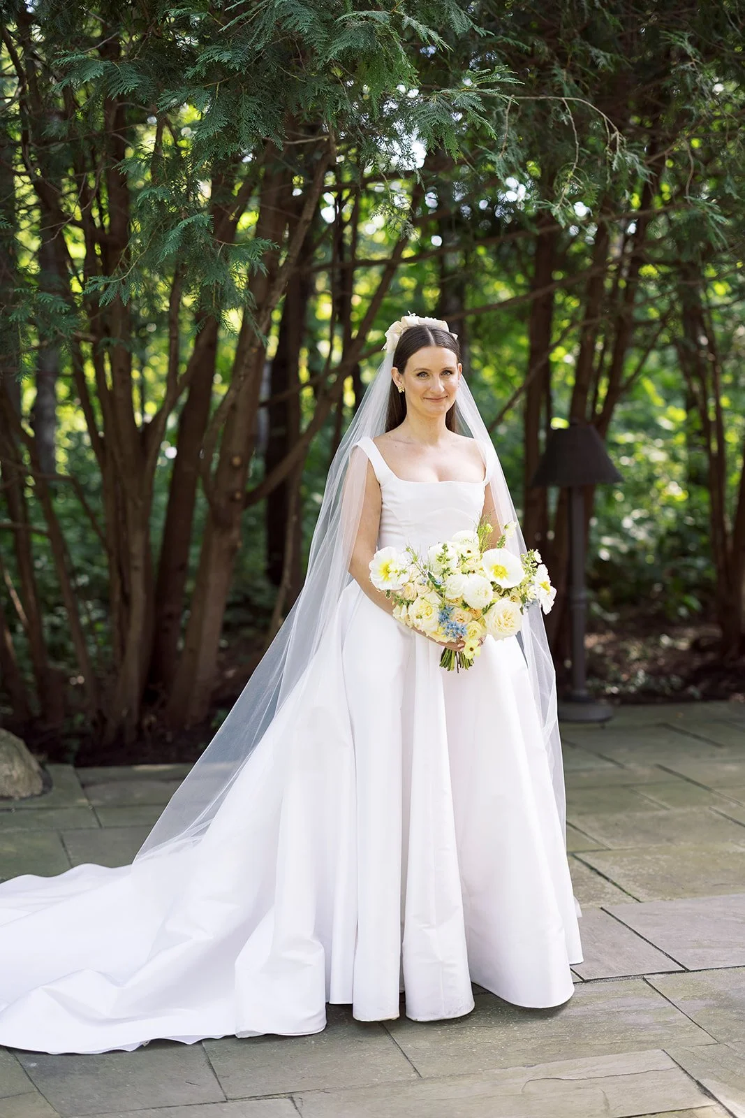 A bride in a white wedding dress holding a bouquet of white and yellow flowers, standing outdoors in a wooded area with green foliage surrounding her.
