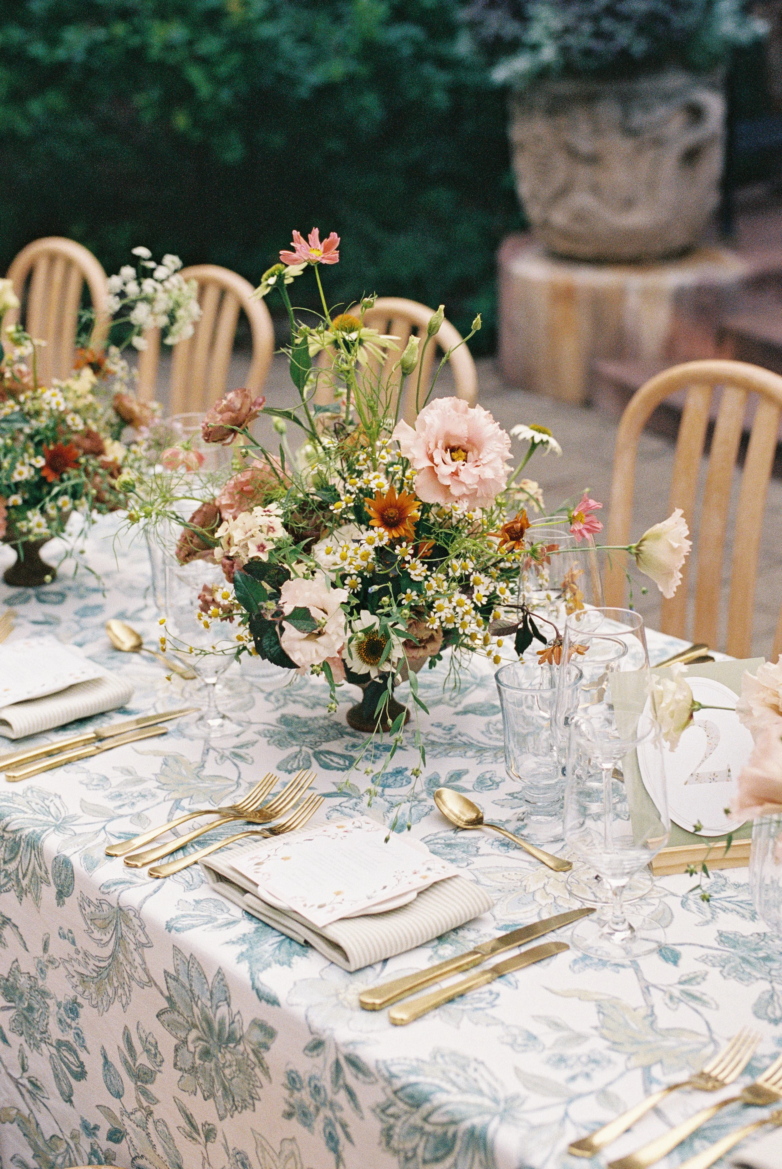 A banquet table set with a floral centerpiece, gold utensils, glassware, and notebooks, outdoors with wooden chairs and a decorative stone element in the background.