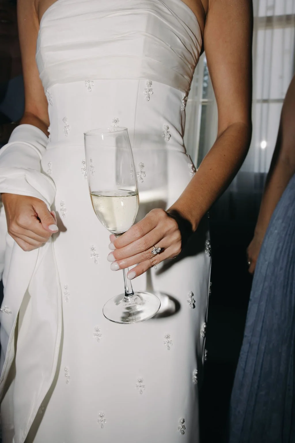 A bride holding a glass of champagne at her wedding, wearing a white wedding dress with beaded embellishments and a diamond ring on her finger.