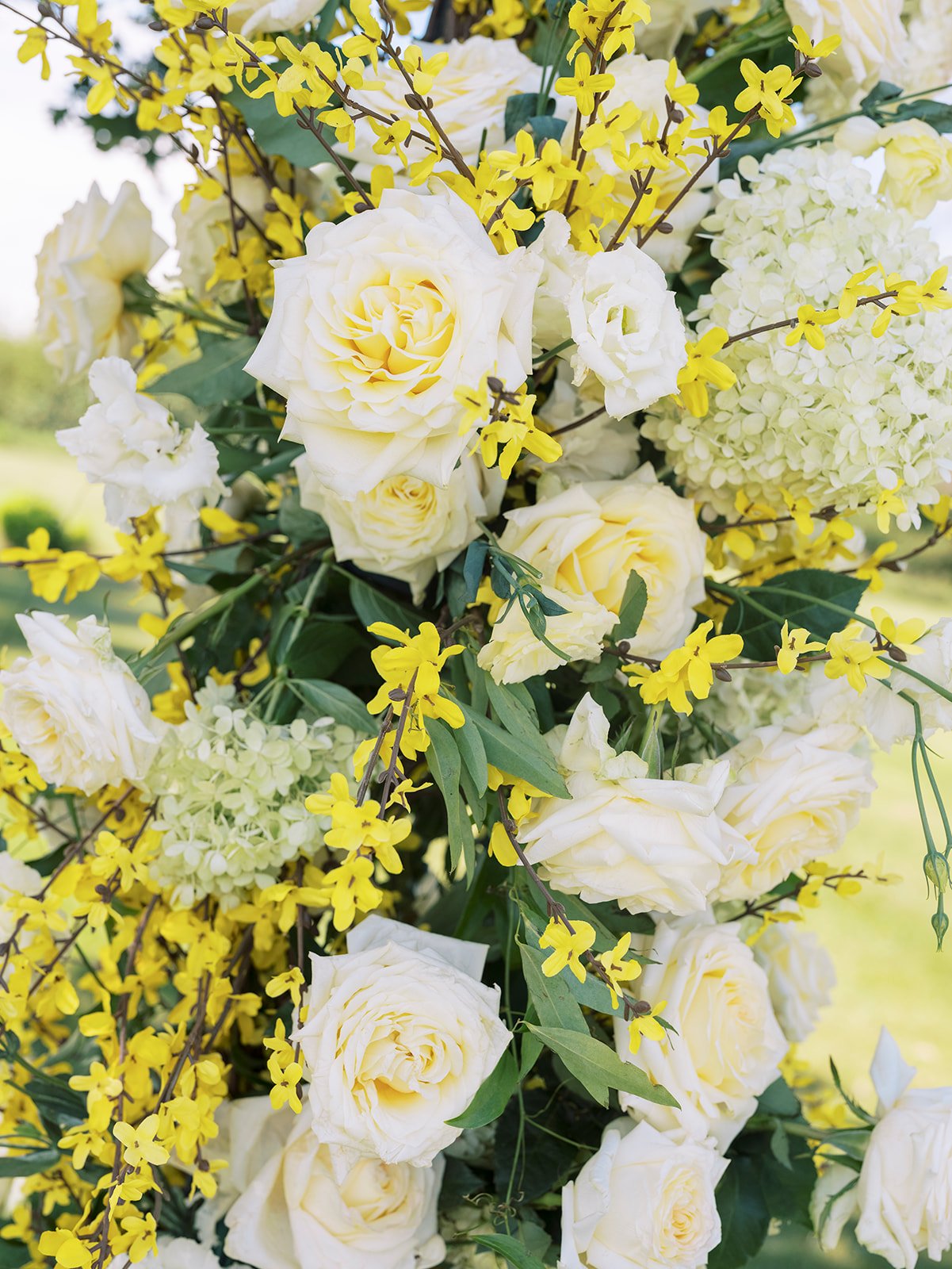 A close-up of a yellow and white floral arrangement with roses, hydrangeas, and small yellow flowers, outdoors on a sunny day.
