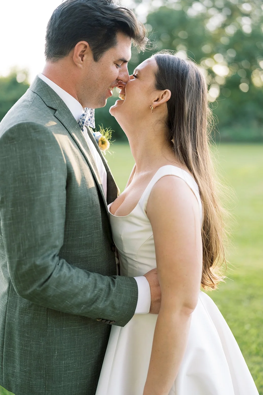 A bride and groom sharing a tender moment outdoors, nose to nose, with blurred green trees in the background.