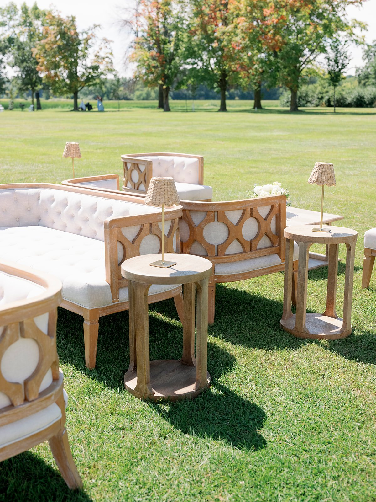 Outdoor furniture arrangement on a grassy field with trees in the background. It includes white cushioned chairs and sofas with wooden frames, and small round wooden side tables with table lamps on them.