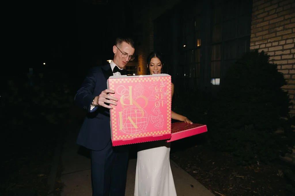 A man in a tuxedo and a woman in a white dress standing outdoors at night, looking at a large pink box with the word 'LOVE' on it.
