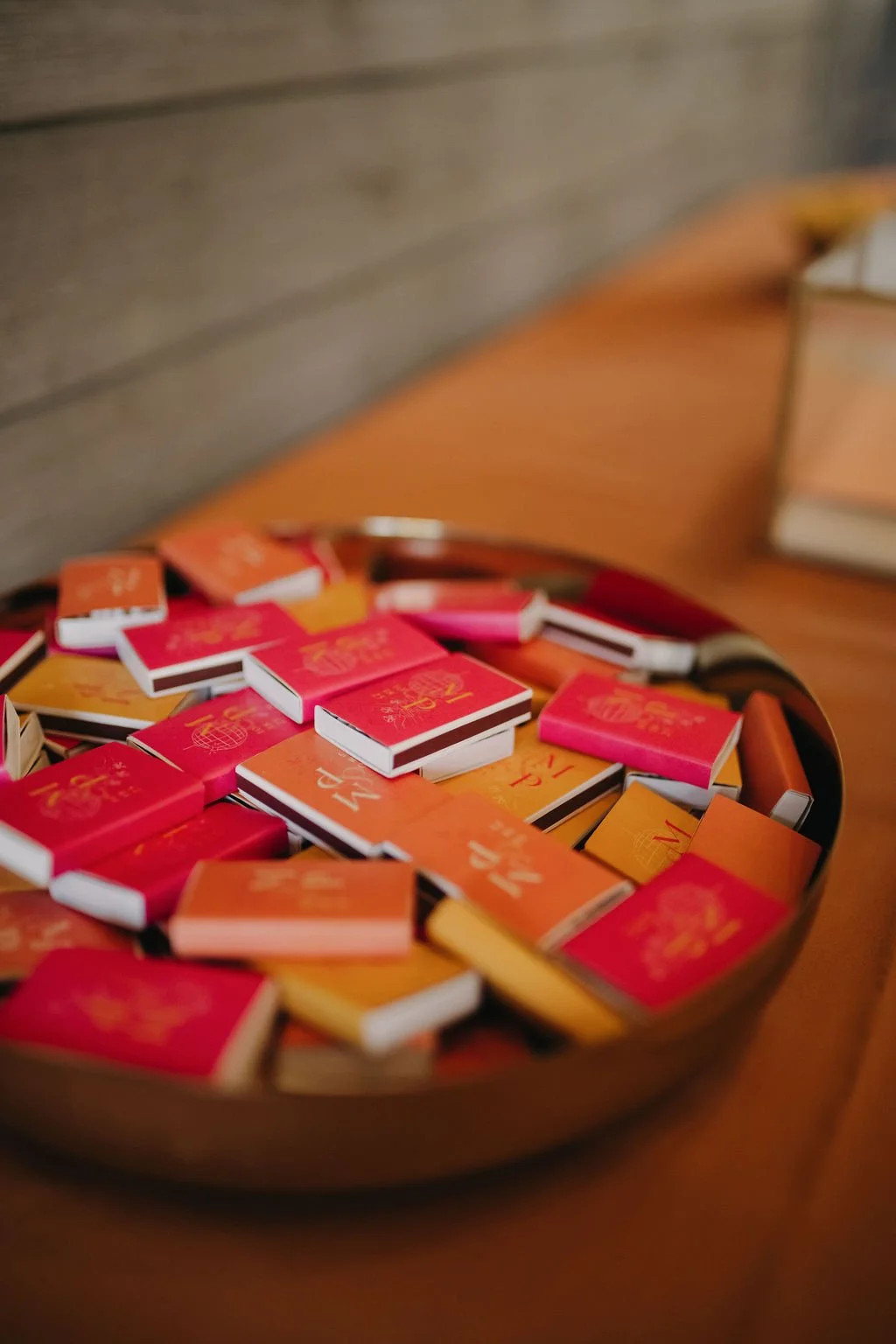 A round metal tray filled with red, orange, and pink matchbooks, placed on a wooden surface with books in the background.