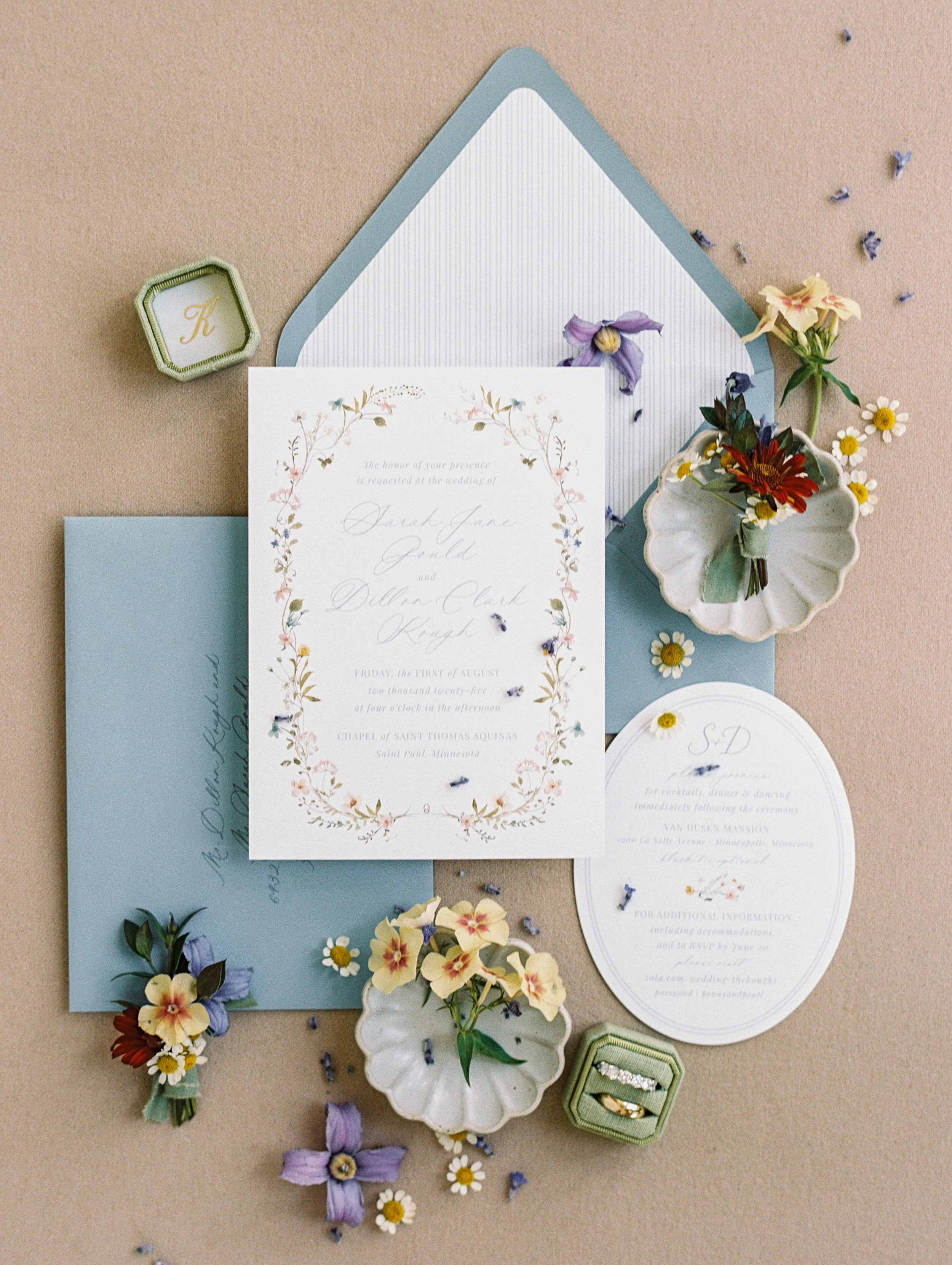 Wedding invitation suite with floral accents on beige background, including a main invite, RSVP card, light blue envelope, floral boutonniere, small dish with flowers, and ring box.