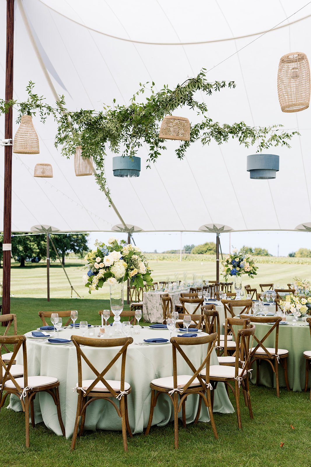 Outdoor banquet setup under a large white tent with round tables covered in white tablecloths, decorated with floral centerpieces and set with glassware and navy blue napkins. Wooden chairs with white cushions surround the tables. Hanging wicker and fabric lanterns and greenery decor are suspended from the tent ceiling.