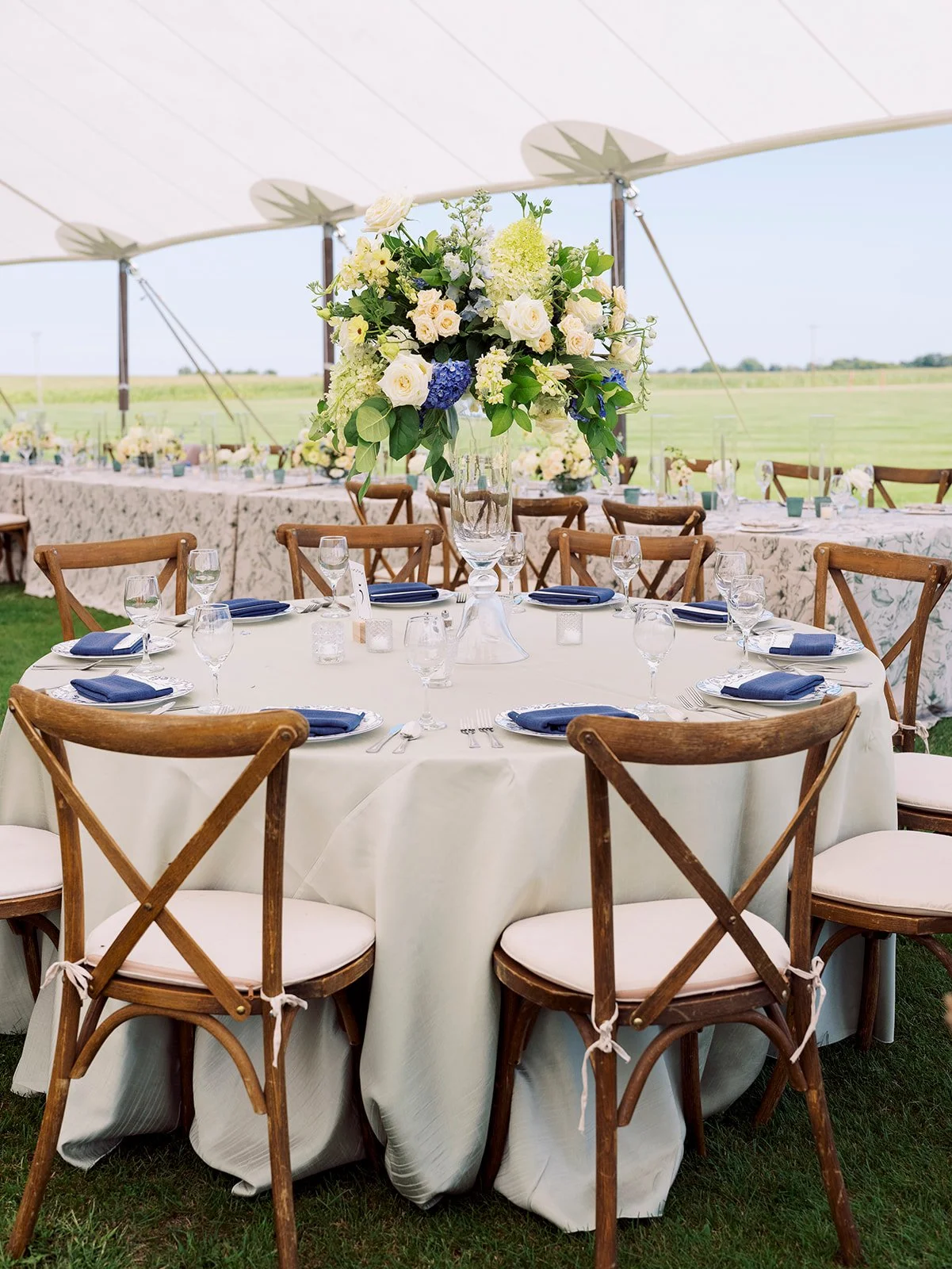 Decorated round banquet table with white tablecloth, navy blue napkins, wine glasses, and silverware, under a white tent. Centerpiece features a tall glass vase with a large arrangement of white and blue flowers, green leaves, and accents. Chairs with wooden frames and white cushions surround the table. Rolling green fields are visible outside the tent.