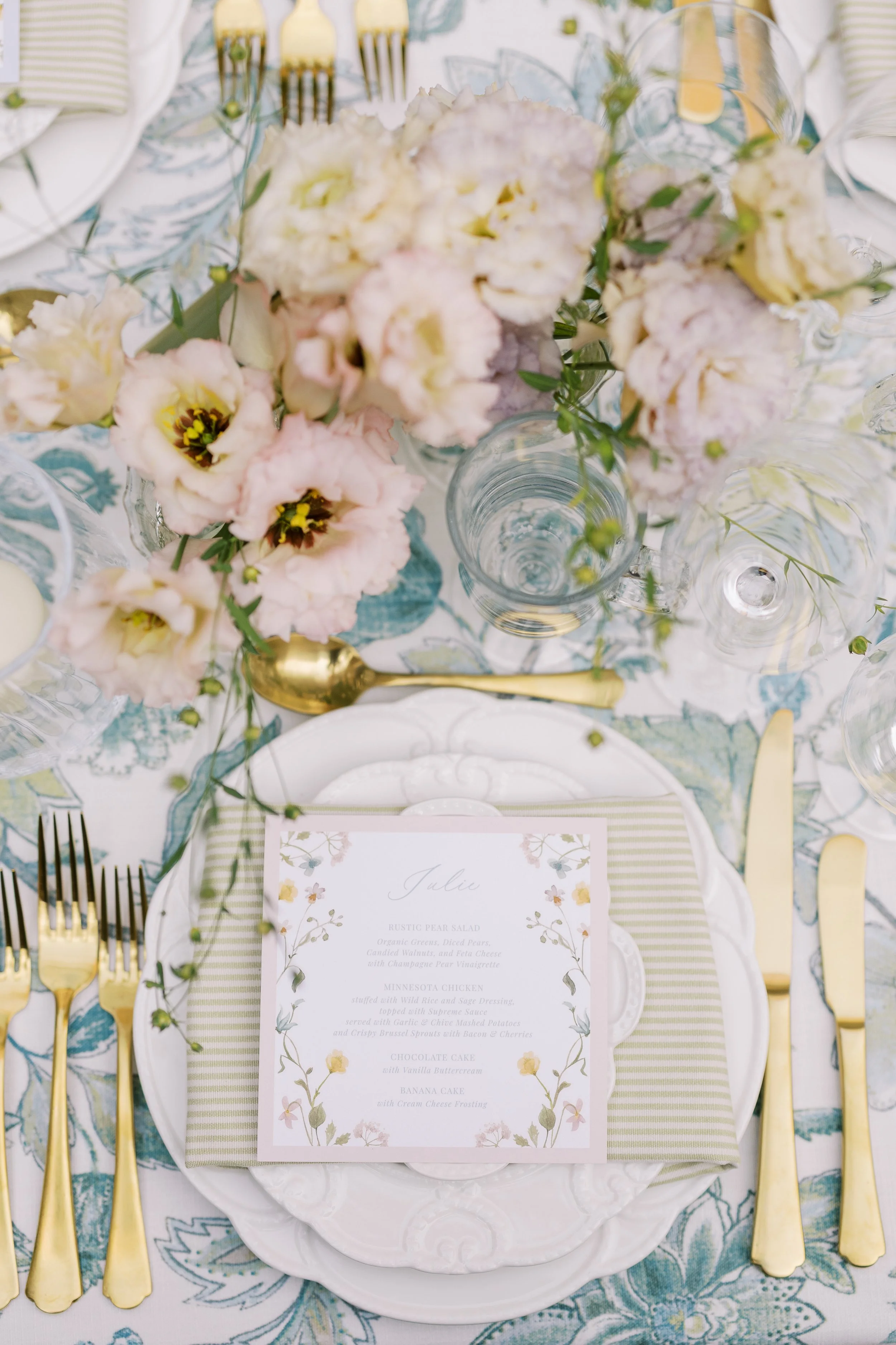 Elegant table setting for a special occasion featuring a floral centerpiece with pink and cream flowers, gold flatware, a decorative menu card with floral border, and a patterned tablecloth with light green, blue, and cream colors.