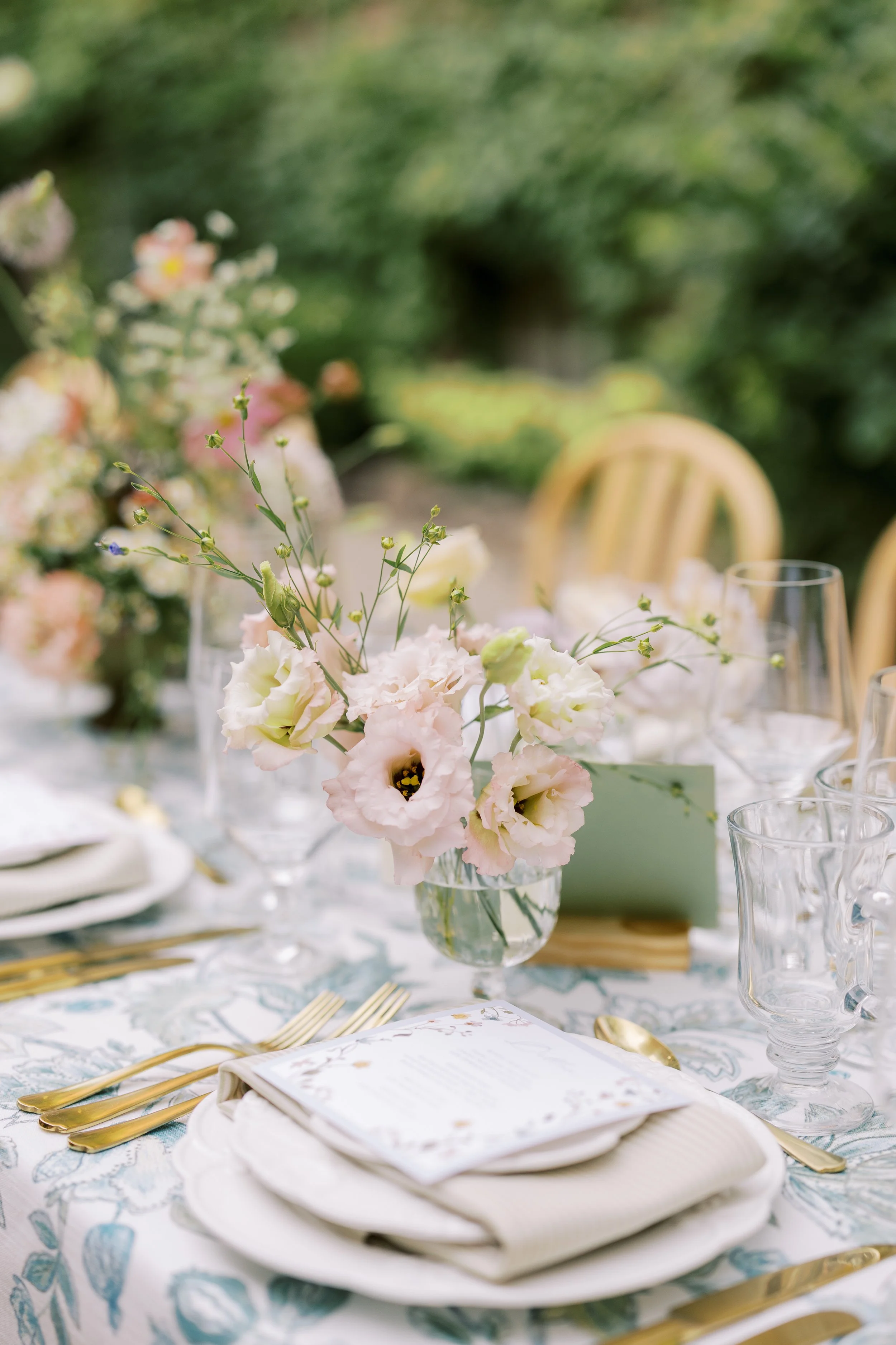 Elegant outdoor table setting with pink and white flowers, clear glassware, gold utensils, and a printed menu.
