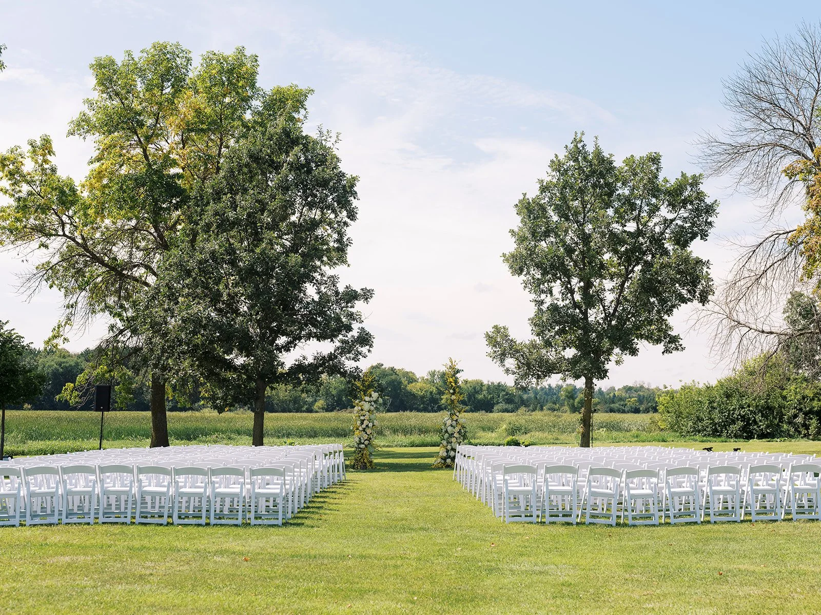 Outdoor wedding setup with white chairs arranged in rows on a grassy lawn, flanked by trees and decorated with floral arches in the background, under a partly cloudy sky.