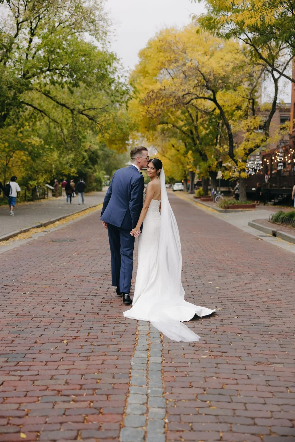 A newlywed couple holding hands on a brick-paved street surrounded by trees with autumn leaves, the bride in a strapless wedding gown and veil, the groom in a blue suit, with people walking and buildings in the background.