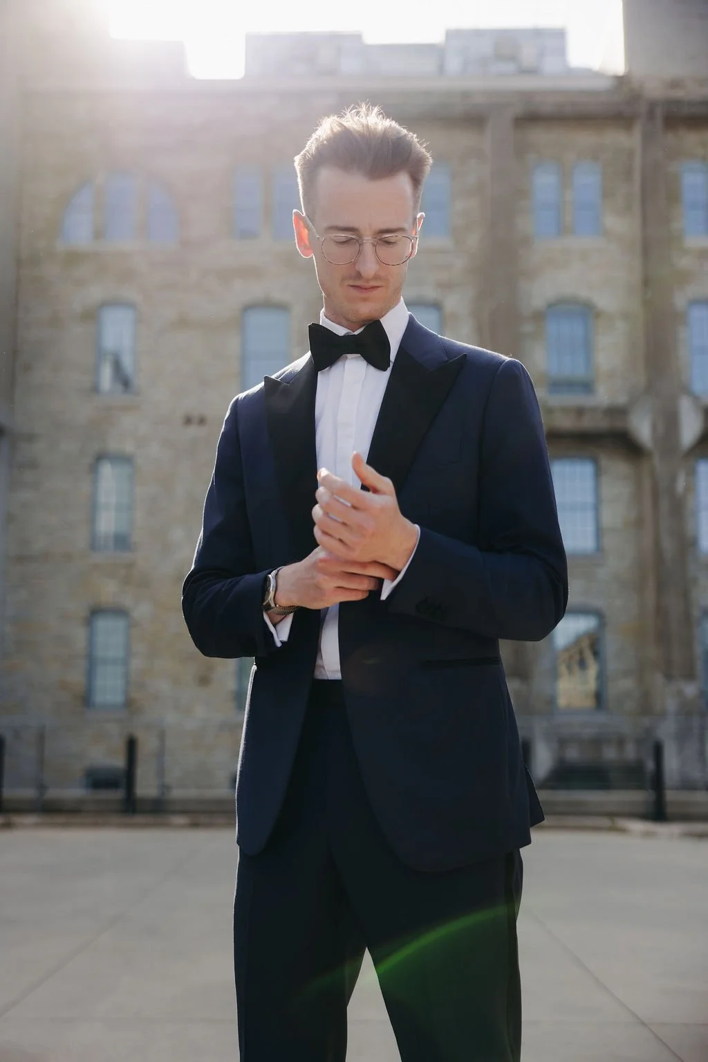 A man in formal tuxedo with black bow tie, glasses, and a wristwatch standing outside in front of an old building with multiple windows, looking down and adjusting his cuff.