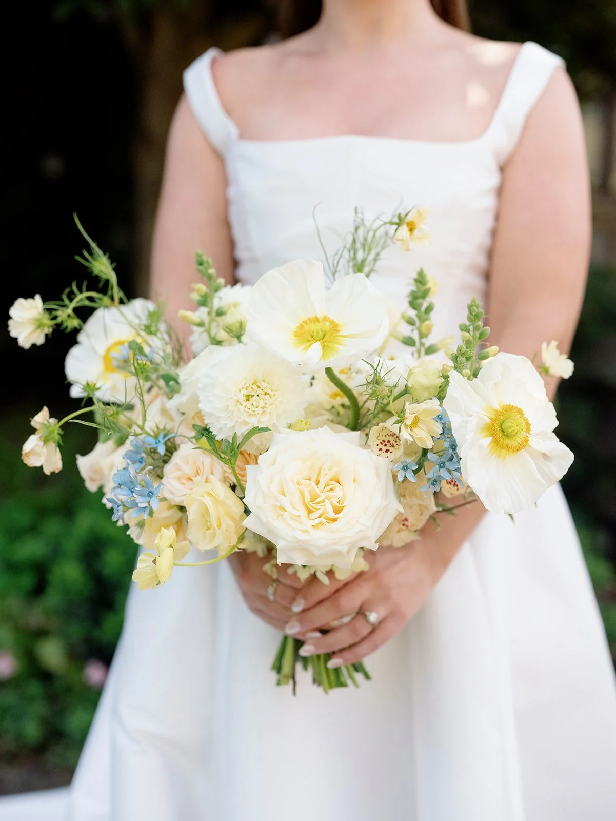 A woman in a white dress holding a bouquet of white and pale yellow flowers, including roses and poppies, with greenery.