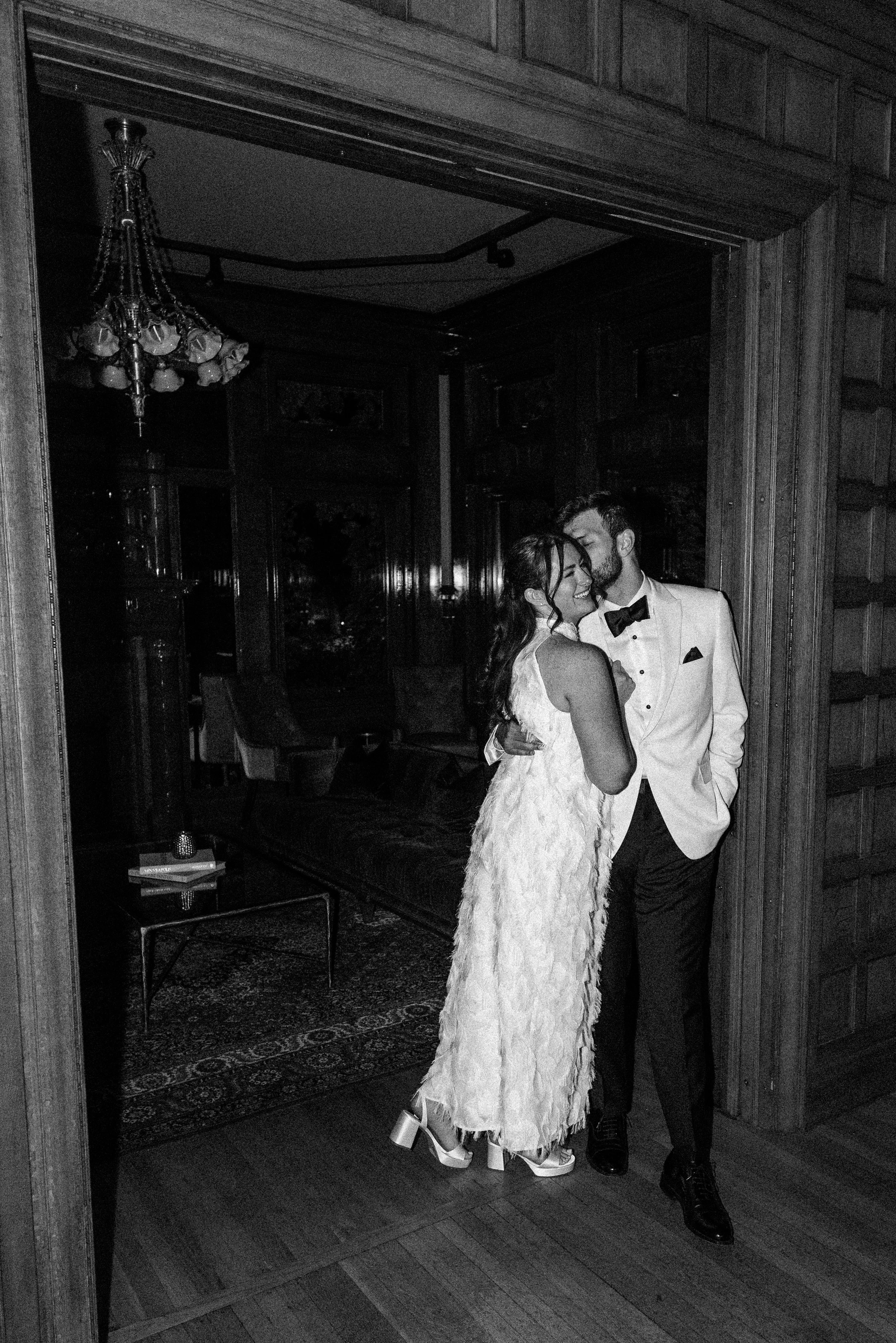 A black and white photo of a couple dressed in formal attire, standing close together in a room with wooden walls and a chandelier, sharing a tender moment.