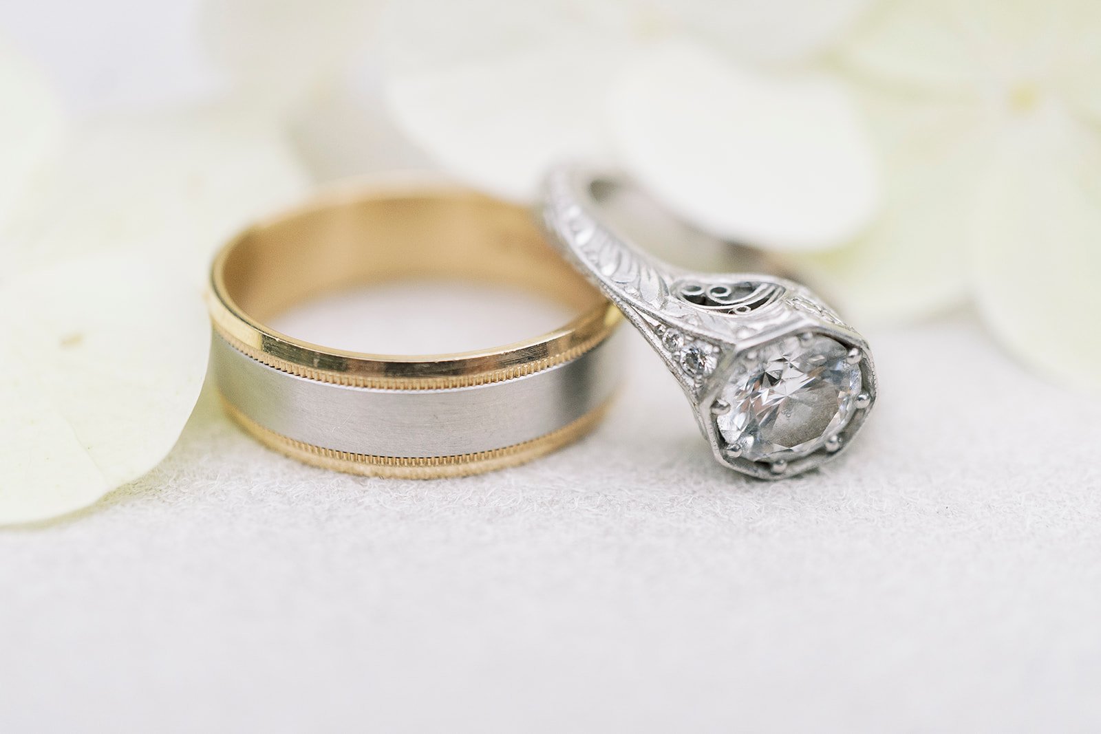 Wedding rings, one gold and silver, and a silver engagement ring with a large gemstone, resting on a white surface with white rose petals.